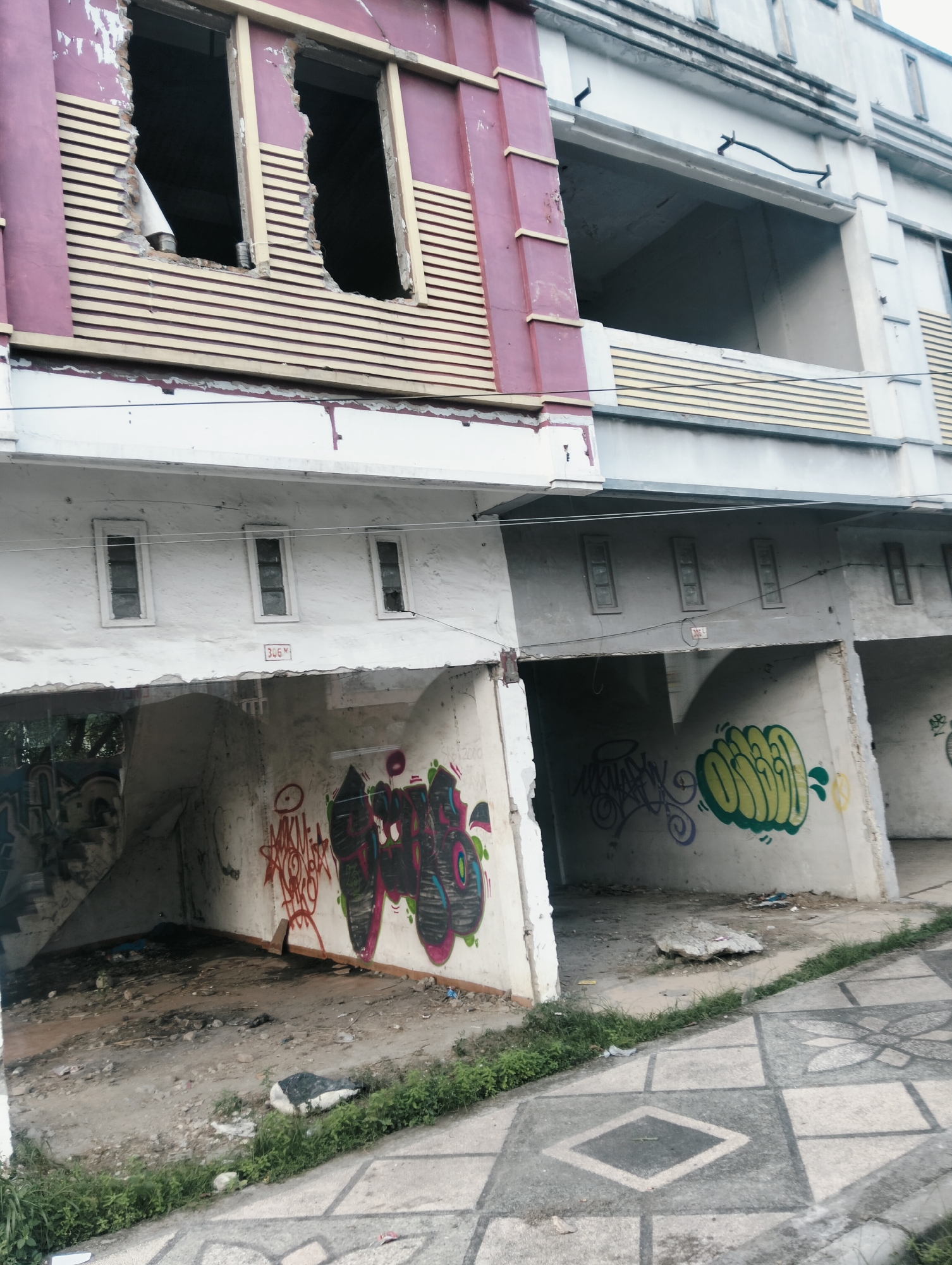 The image depicts the exterior of a derelict building in the city of Medan, Indonesia. The building appears to be several stories high, with sections of the upper facade showing exposed brick and broken windows. The lower level is open, resembling a parking or storage area, with walls covered in colorful graffiti. The ground in front of the building is paved with a decorative pattern of squares and diamonds, and there is some overgrown grass and debris. The scene conveys a sense of abandonment and decay.