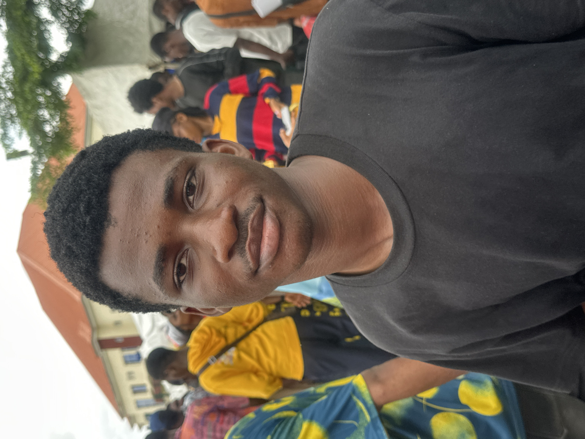 A medium close-up shot shows a young Black male facing forward, his head and shoulders filling the foreground. He has short, dark, curly hair and dark skin. His expression is neutral, with his eyes looking directly at the camera and a slight mustache visible above his upper lip. He is wearing a plain black t-shirt.
In the blurred background, a crowd of people is visible. To the left, a building with an orange roof and yellow walls stands under a cloudy sky. Several individuals are walking and interacting within the space. One person in the mid-ground wears a bright yellow hooded sweatshirt and dark pants, with a dark bag slung across their body. Another person to their right wears a horizontally striped long-sleeved shirt in red, yellow, and dark blue. The overall environment suggests an outdoor gathering or public space in Ihiagwa, Nigeria.