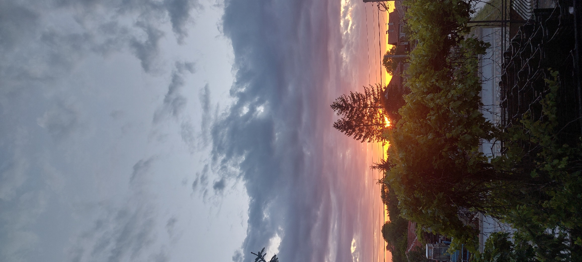 The image shows a view of Yagodovo, Bulgaria at sunset. Dark clouds fill the left portion of the sky, while shades of orange and pink sunset light dominate the right. Silhouetted trees are in the center, with additional vegetation and a building on the right. Power lines are visible, crossing the scene horizontally. A fence is present on the far right.