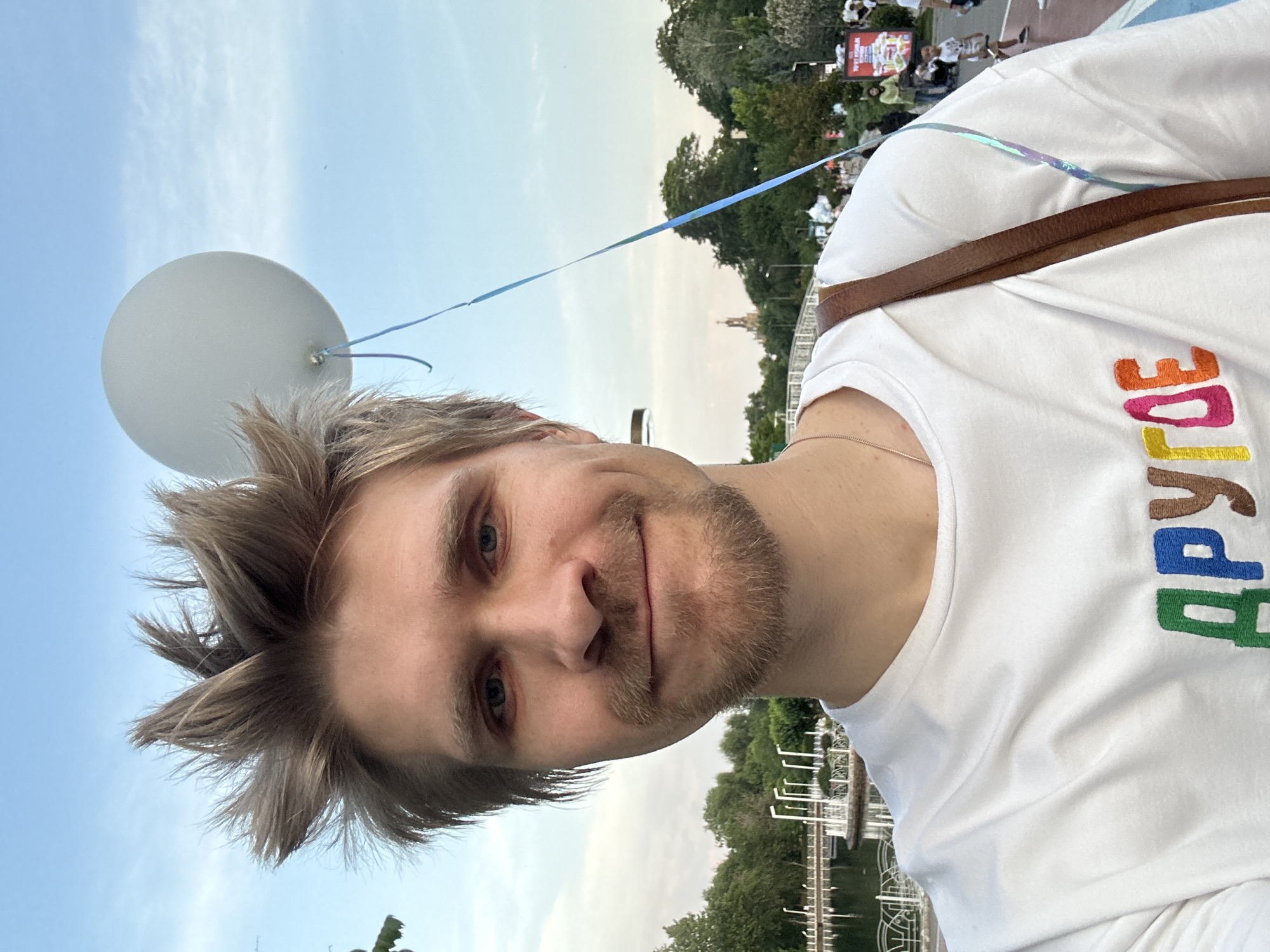 A selfie shows a man holding a white balloon on a string, posing in a public place near a pond and trees. He is wearing a white t-shirt with a word in Russian embroidered in rainbow colors. The man has short, light brown hair and a beard. A brown strap is visible across his shoulder. In the background, a cityscape can be seen. Based on available information, this could be in Tashkent, Uzbekistan. Other people are visible in the distance.
