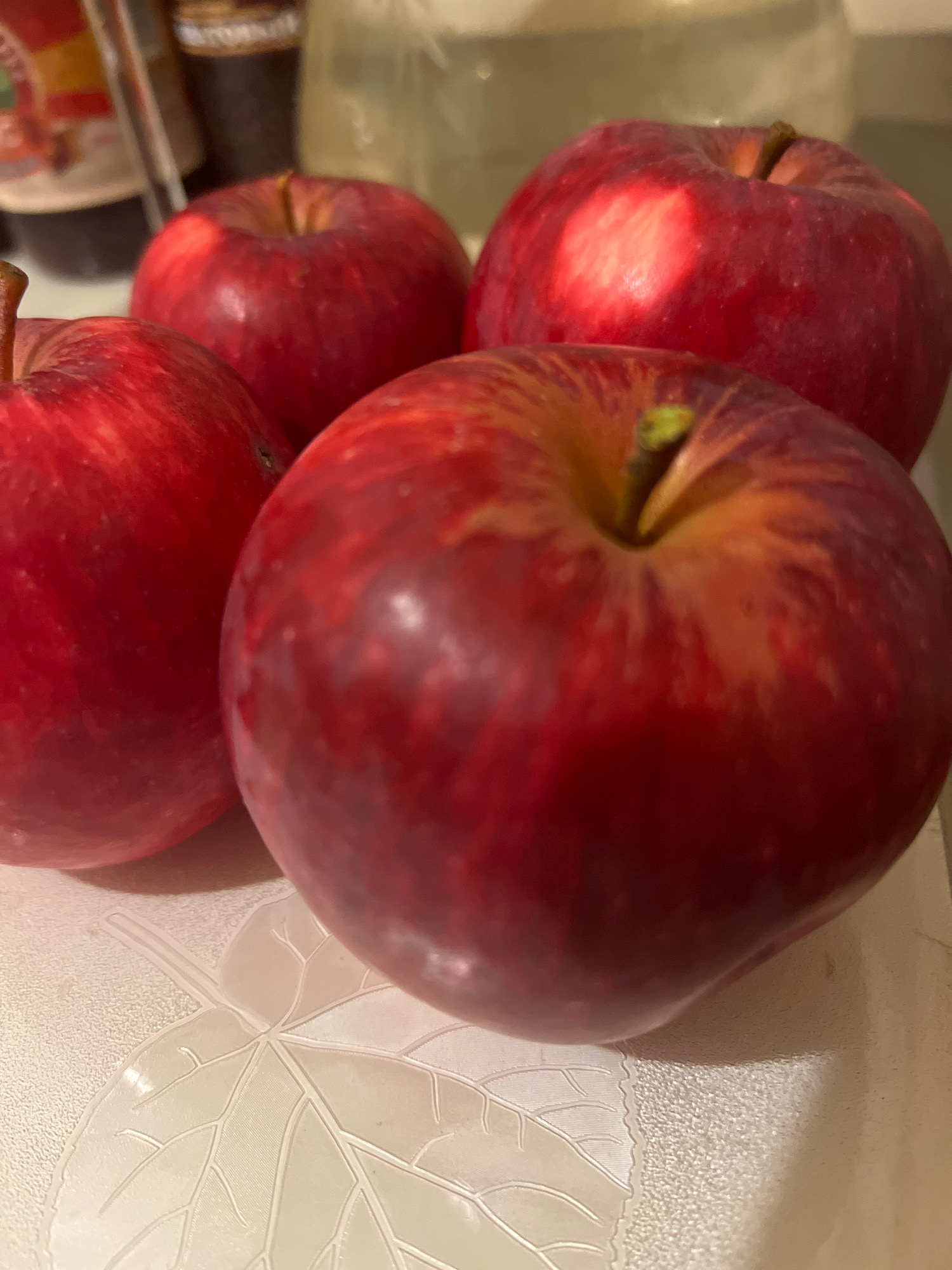 Four red apples are arranged on a textured, light-colored surface. The apples are round and vary slightly in size, with visible stems. The surface has an embossed pattern resembling leaves and veins. In the background, out of focus, are bottles and a blurred glass object. The lighting creates highlights and shadows on the apples.