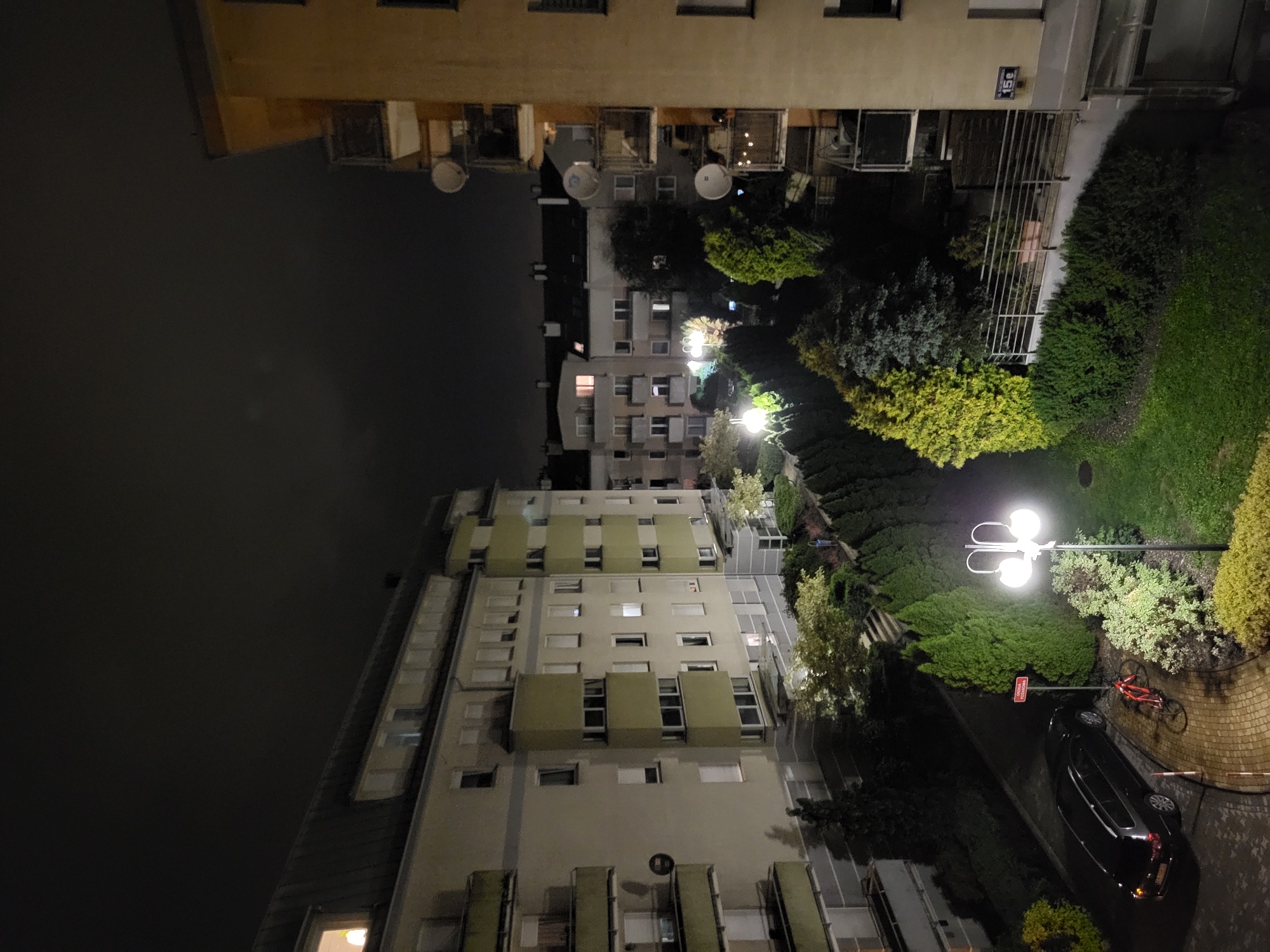 This is an eye-level, medium shot of a residential area at night. The scene is illuminated by streetlights and lights from within apartment buildings. On the left is a large, multi-story apartment building with a dark roof and a light-colored facade. Several windows are illuminated, indicating occupancy. Balconies with railings are visible on the building. In the foreground, a dark-colored station wagon is parked on a cobblestone driveway. A red bicycle is leaning against a signpost next to the car. A street lamp with two white globes stands to the right of the driveway, illuminating the surrounding greenery. A second, taller apartment building, primarily yellow, is visible on the right side of the frame, also featuring balconies and satellite dishes. Trees and shrubs are planted between the buildings, creating a landscaped courtyard area. The sky is dark and overcast.