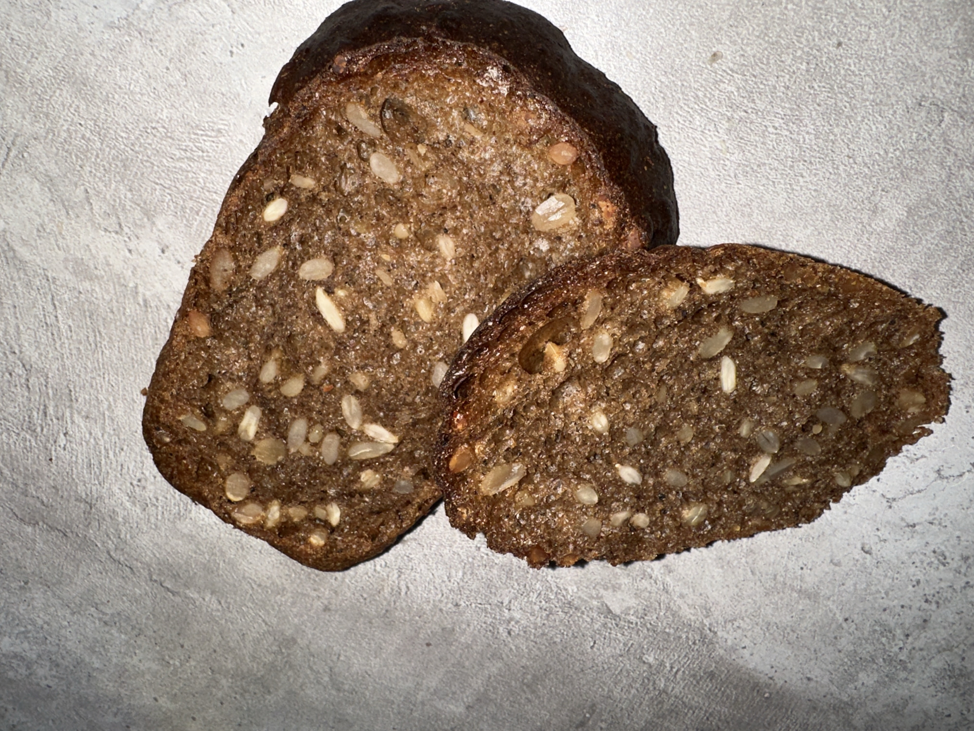 The image shows two slices of dark, seeded bread on a gray, textured surface. The larger slice is partially obscuring the smaller one. The location of the image is not discernible and cannot be determined as Chaikovsky, Russia. There are no people in the image. The bread slices are the primary subjects. No actions or interactions are visible. The environment consists of the bread and the underlying surface.