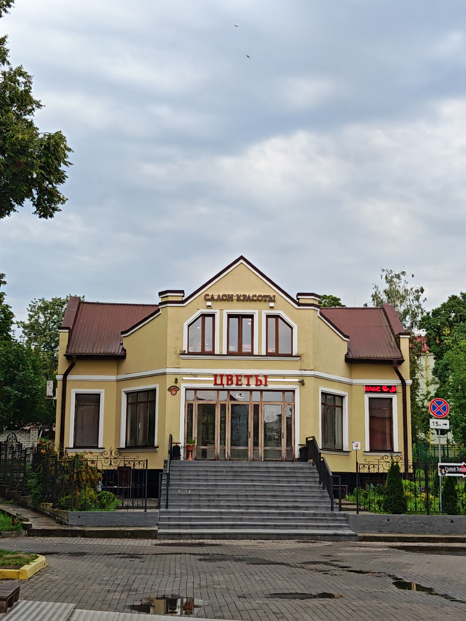 The image depicts a yellow, two-story building with a brown, sloped roof. The building features several large windows, some with arched tops, and a set of wide stairs leading up to a double-door entrance. Red neon signs on the building read "ЦВЕТЫ" (Flowers) and "САЛОН КРАСОТЫ" (Beauty Salon), and "КАФЕ" (Cafe) with a paw print graphic. The building is situated on a paved street with some puddles reflecting the overcast sky. Trees and a black metal fence surround the property. A red and blue traffic sign with a white circle and diagonal line is visible on the right side of the image, indicating a no parking zone, and a directional sign with Cyrillic text points to the right. The scene appears to be in a city, with hints of urban infrastructure like streetlights and signage. The overall atmosphere is muted due to the cloudy weather. The building is identified as located in Hrodna, Belarus.
