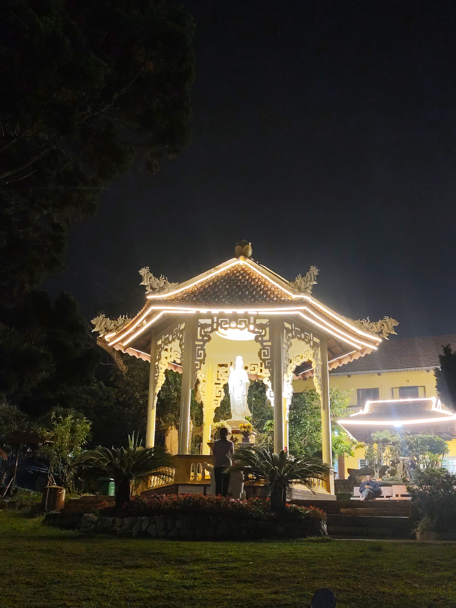 The image presents a serene nighttime scene, likely within a spiritual complex or temple garden in Da Lat, Vietnam. A prominently featured octagonal pavilion stands centrally, its ornate, multi-tiered tiled roof and intricately carved supports brilliantly illuminated by warm string lights. Dragon figures adorn the roof edges, characteristic of traditional Vietnamese architecture.
Inside the pavilion, a tall, white statue, possibly of a deity such as Guan Yin or a Bodhisattva, is brightly lit, serving as the focal point. One person stands before this statue, facing it in what appears to be an act of reverence or contemplation. To the right and further back, another individual is seated on a step or bench, seemingly engaged with a mobile phone.
The pavilion is surrounded by a well-maintained garden area, featuring a grassy foreground, circular flowerbeds with red and green foliage, and small palm-like plants. Beyond the pavilion, a multi-story yellow building with a dark tiled roof and windows is visible, also illuminated, suggesting it is part of the same complex. Dark, dense trees frame the top and left edges of the frame, silhouetted against the deep black night sky. The overall impression is one of peaceful solemnity, enhanced by the soft, artificial lighting against the darkness. No visible text is present in the image.