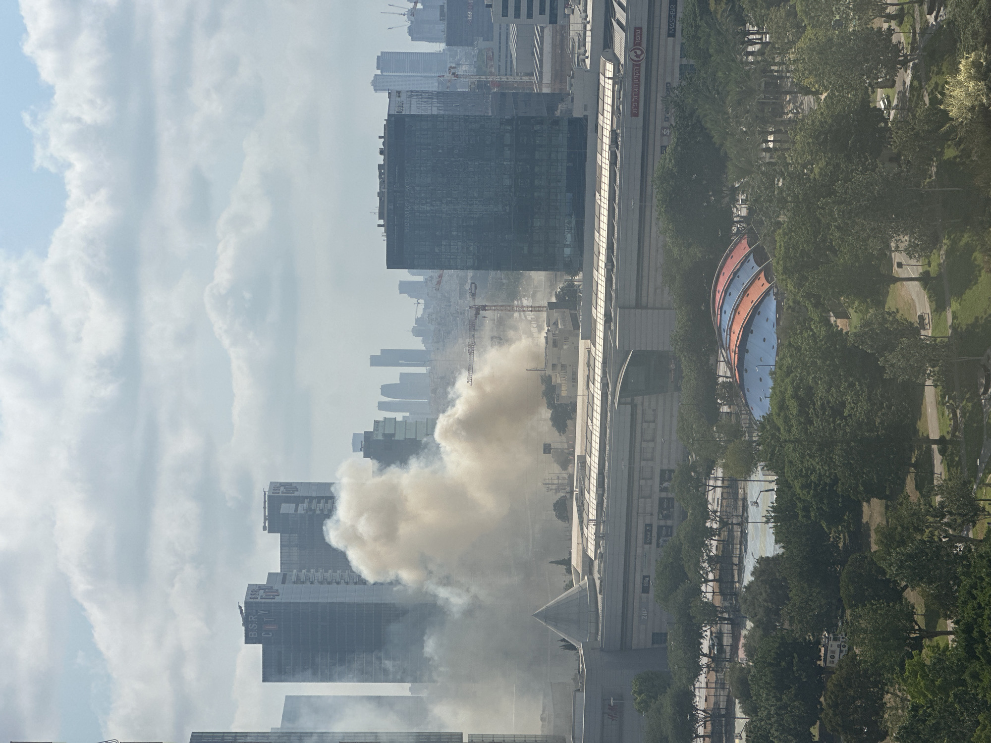 The aerial view captures a section of the city of Petah Tikva, Israel, dominated by urban architecture and a plume of smoke. In the background, several high-rise buildings, including the B.S.R City complex, punctuate the skyline under a sky filled with clouds. A prominent, multi-story building labeled "HOLMES PLACE" is visible in the mid-ground. The central focus of the image is a large, dense cloud of yellowish-white smoke billowing from behind a large, modern shopping center. This shopping center, featuring a pyramidal structure and visible signage for brands like H&M, ZIP, and BIGA, is located in the foreground. Beyond the shopping center, a park area is visible, with sports courts and a distinctive dome-shaped structure with red and blue panels. Trees and greenery surround the park and shopping center. There are no visible people in the frame, but there is an ambulance present in the park area. Several construction cranes are also scattered throughout the scene, suggesting ongoing development.