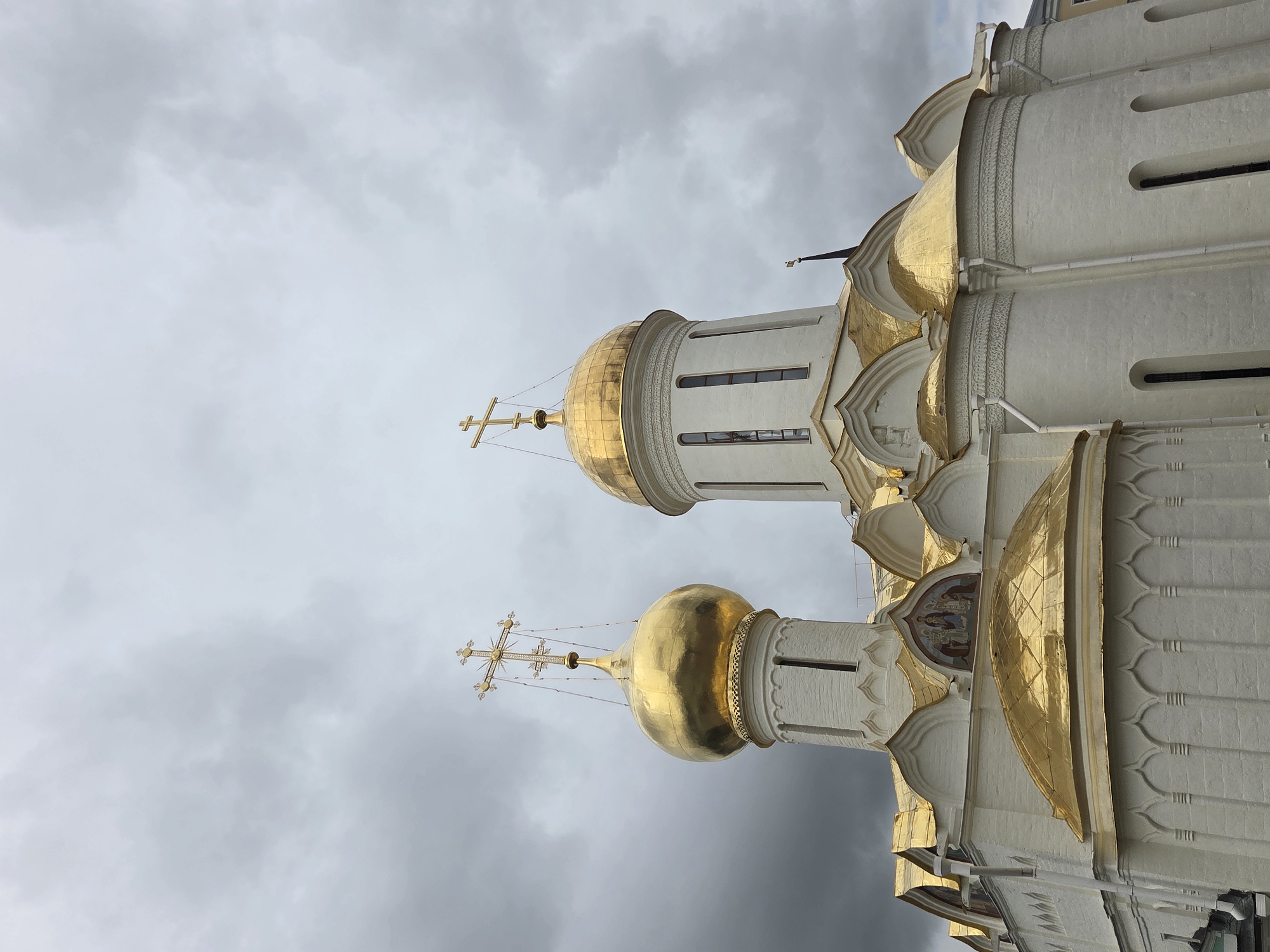 This media file displays a close-up, low-angle view of the white and gold domes of a Russian Orthodox church against a cloudy sky. The architecture features intricate detailing, with multiple golden onion domes topped with ornate crosses. The church walls are white with decorative architectural elements, and narrow windows are visible. The sky is overcast with dark, heavy clouds, suggesting it might be a gloomy or stormy day. The image is oriented vertically, with the top of the church appearing on the right side. There are no people visible in the image. The scene appears to be Sergiyev Posad, Russia, known for its religious sites.