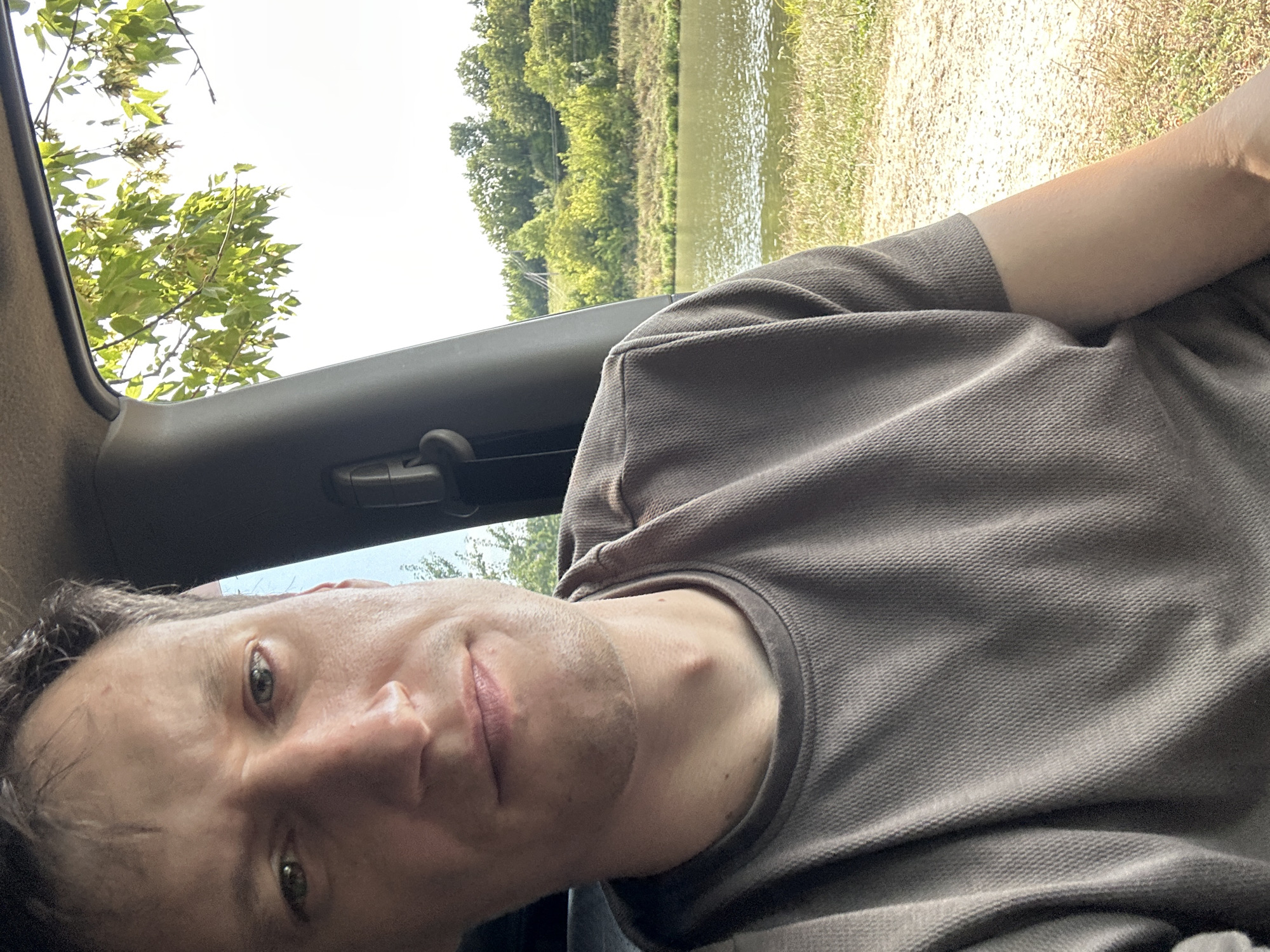 A medium shot captures a man in the driver's seat of a vehicle, looking towards the camera. He is wearing a grey, textured polo shirt. The background visible through the car window shows a body of water, a grassy bank, and a treeline against a bright sky. The image is likely taken on a sunny day. The context of Makiv, Ukraine is not discernible from the visual information provided.