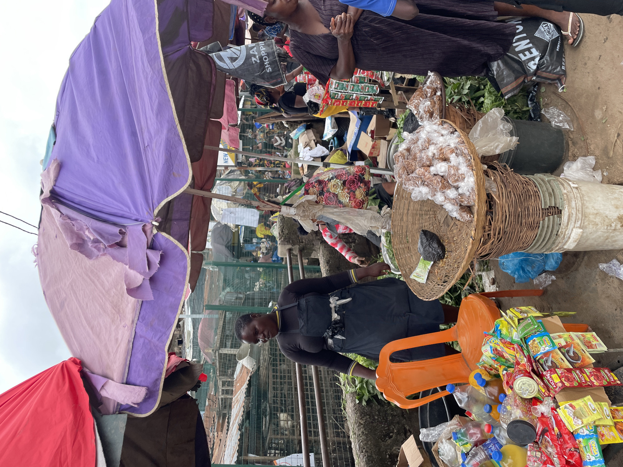 A marketplace scene in an outdoor setting features several people and numerous goods for sale. In the foreground, a woman wearing a black long-sleeved shirt and a dark apron stands behind a table laden with various food items and packaged goods, including bottles of liquids, cans, and small packets of what appear to be spices or seasonings. An orange plastic chair is placed behind her. To the right, another woman in a dark purple dress is visible, and beside her is a large woven basket filled with small, individually wrapped items. In the background, more stalls and people are present, along with caged structures. Overhead, multiple colorful umbrellas, predominantly purple and red, provide shade, with some appearing worn and damaged. The ground is dirt and concrete.