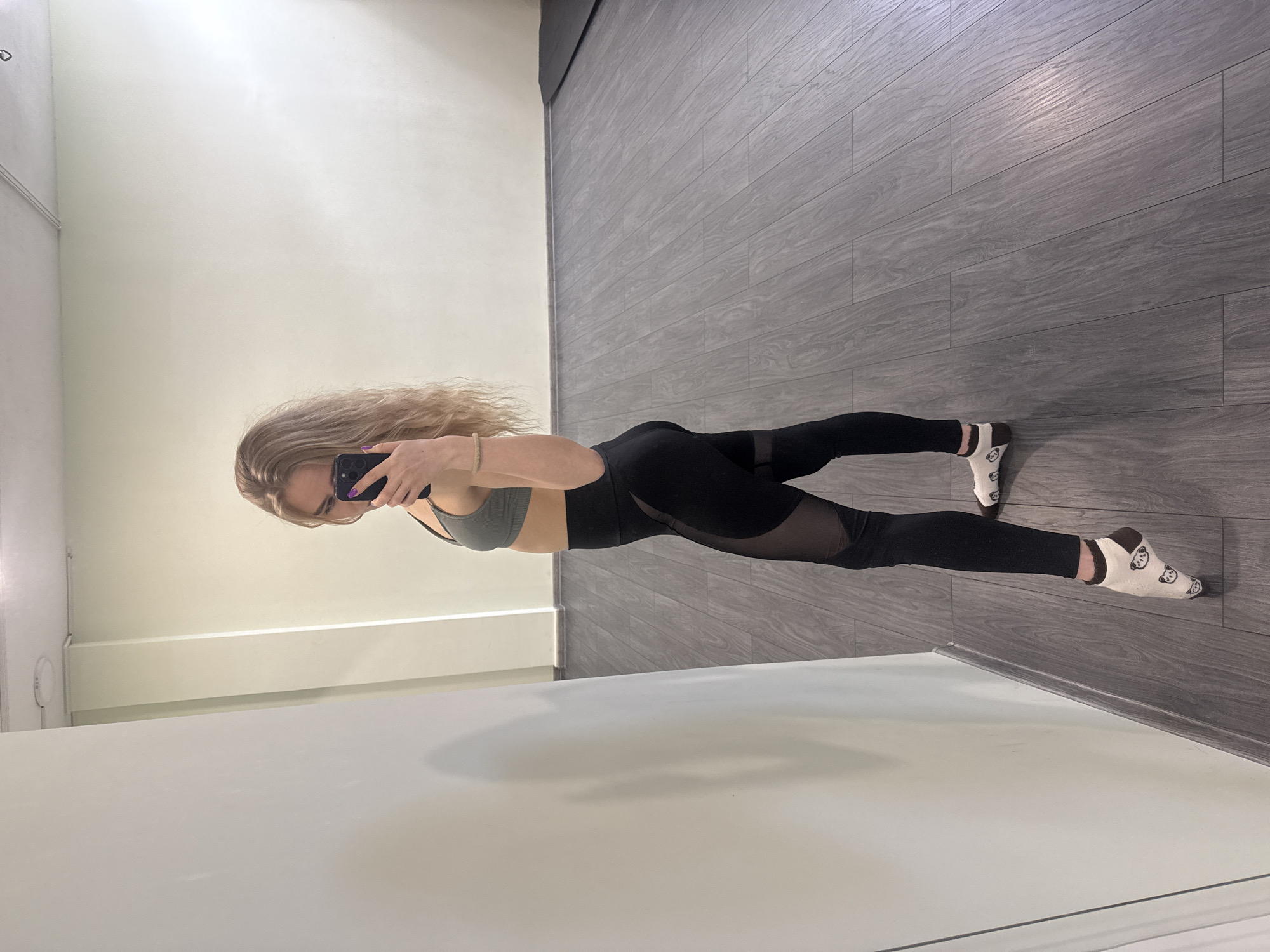 A young adult woman stands in front of a mirror, taking a selfie with a dark-colored smartphone that obscures part of her face. She has long, wavy blonde hair and wears a dark olive green ribbed sports bra paired with black leggings featuring sheer mesh panels on the thighs. A light-colored beaded bracelet is visible on her wrist, and her fingernails are painted purple. She is wearing light-colored socks with dark brown cuffs and small, cartoon-like animal faces printed on them.
The setting appears to be indoors, likely a gym, dance studio, or a room with a large mirror. The background consists of a plain light cream or beige wall on one side, and a dark grey wood-look paneled wall on the other. The floor is also dark grey with a wood grain pattern. The lighting is artificial, suggesting an indoor environment, with some reflections visible on the floor and mirror surface. The woman's posture is casual as she holds the phone to capture her reflection. No text is visible in the image.