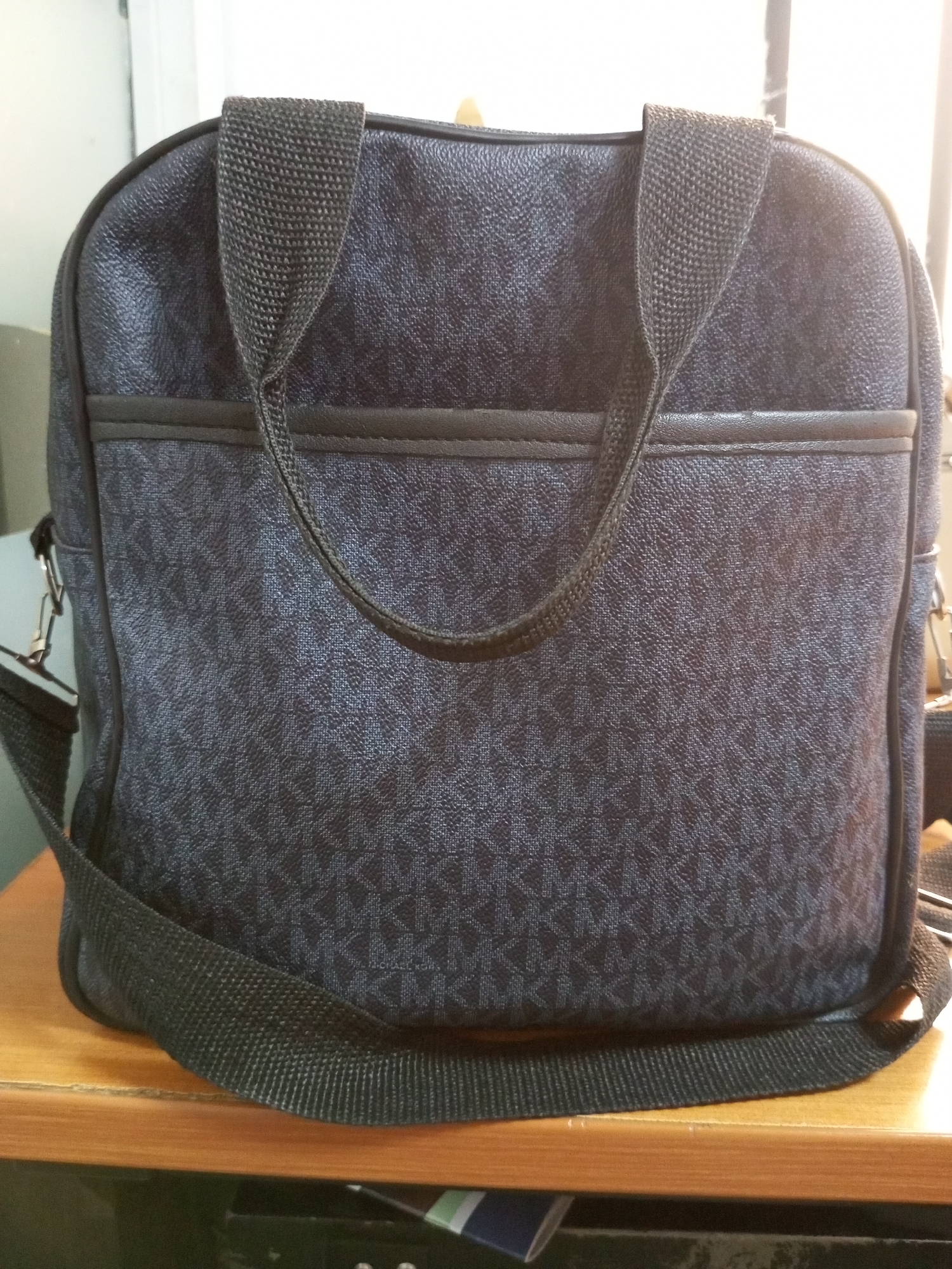 A dark, patterned bag is displayed on a wooden surface. The bag has a repeating geometric pattern in a slightly lighter shade of dark blue or black. It features two dark fabric handles and a long, adjustable dark fabric strap attached to metal clasps on the sides. There is a horizontal pocket across the front of the bag, outlined with dark trim. The background is out of focus, showing what appears to be a light-colored wall and a glimpse of a window with natural light coming through, suggesting it is daytime. The scene is indoors, and the lighting creates a slight glare on the bag. The overall impression is a simple product shot or a photograph taken in a domestic or casual setting, likely in Bukuru, Nigeria, given the location context. There is no visible text on the bag or in the immediate surroundings.