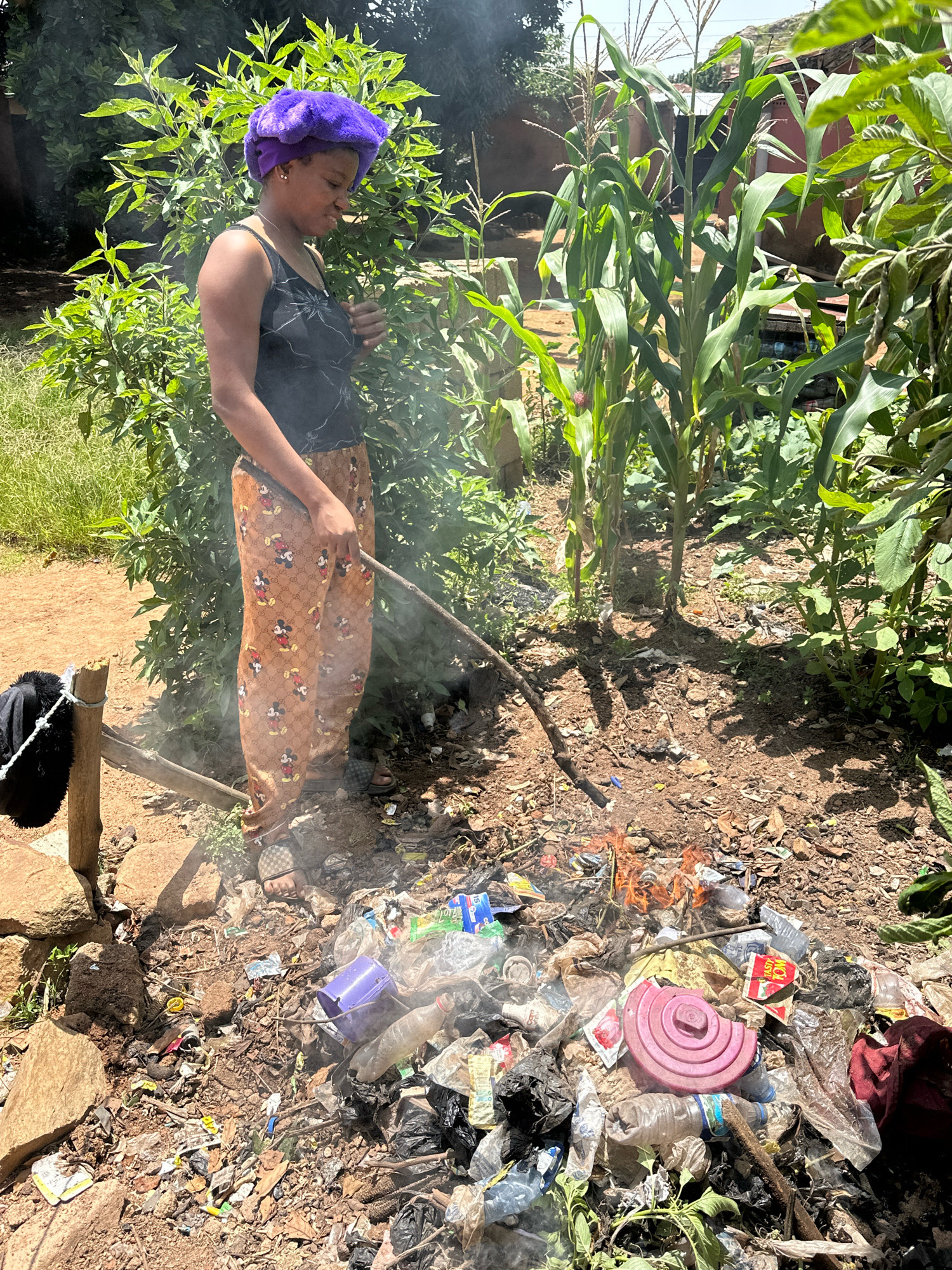 In Gura Top, Nigeria, a young woman wearing a purple headscarf and patterned pants with images of Mickey Mouse is tending to a small, smoldering trash fire. She holds a stick, using it to poke at the burning debris, which includes plastic bottles, bags, and other assorted waste. The fire is situated on uneven, dusty ground littered with more trash. Behind her, lush green foliage and stalks of corn grow, suggesting an outdoor, perhaps semi-rural, setting. A wooden post with a black item hanging from it is visible to her left. The overall scene depicts an act of waste disposal in a somewhat neglected environment.