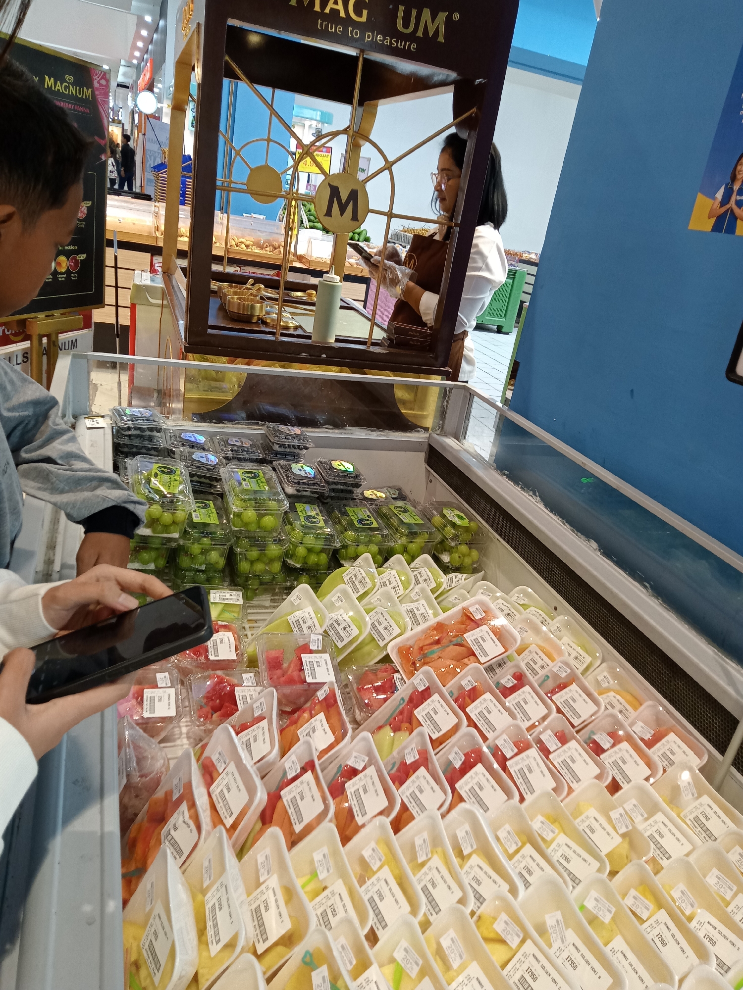 In the city of Batu, Indonesia, at a retail establishment, a person is holding a smartphone and appears to be taking a photograph or recording video. They are standing in front of a refrigerated display case filled with various pre-packaged fruits, including grapes, blueberries, and sliced fruits like watermelon and cantaloupe. Behind the display case, a woman wearing glasses and an apron is visible, attending to a food preparation area. The display case itself is part of a larger market or store, with signage and other retail elements visible in the background. The overall scene captures a moment of shopping or browsing within a grocery store environment.