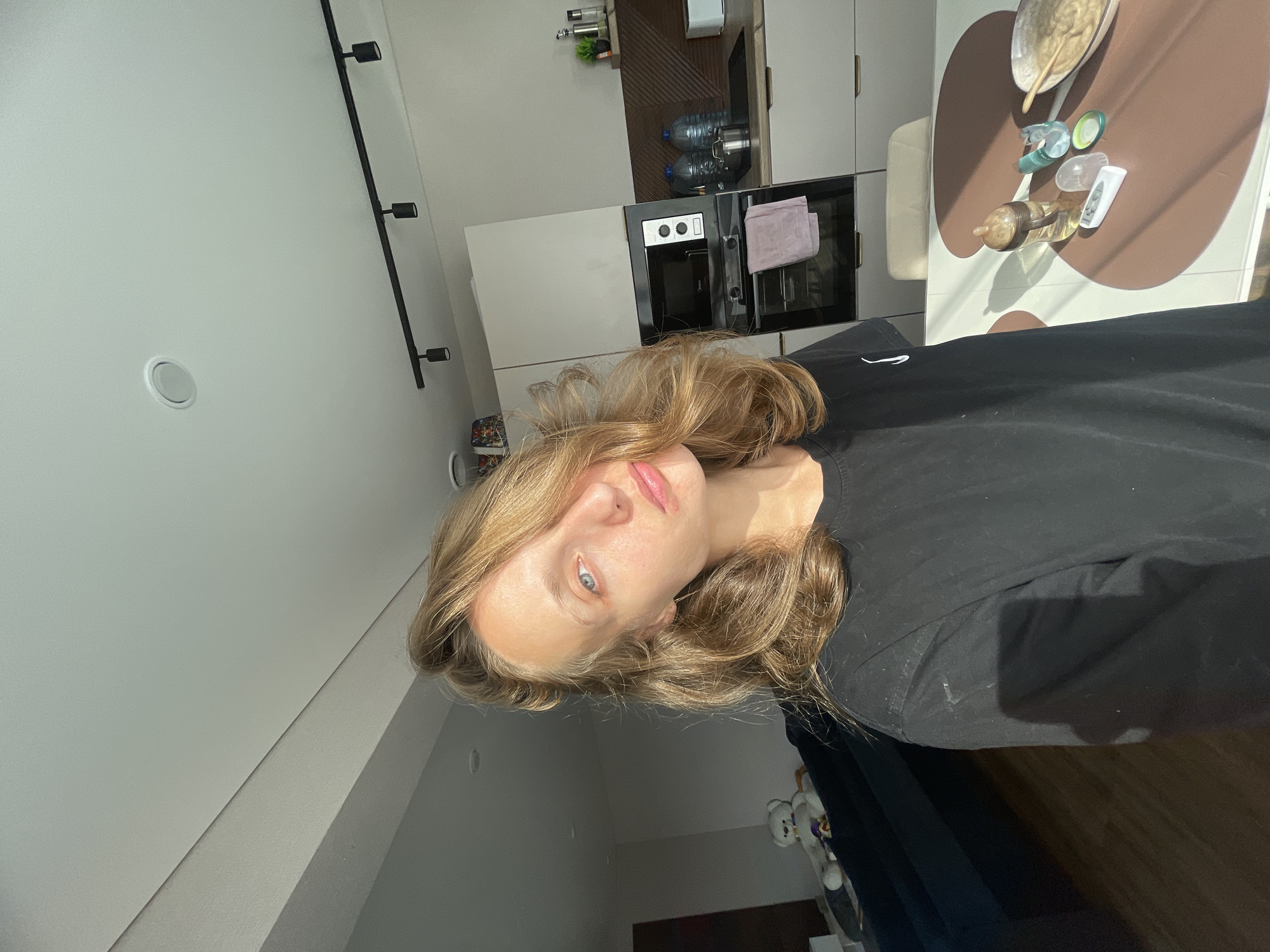 A medium-shot, eye-level, selfie-style photograph captures a young woman with blonde hair, fair skin, and blue eyes. She is wearing a black t-shirt and is positioned in the foreground. Her head is slightly tilted, and her hair partially obscures her face. Sunlight streams in from the right side of the frame, illuminating her face and creating shadows.
The background reveals a domestic interior, likely a kitchen and living area. To the left, a dark blue sofa is visible with a white stuffed teddy bear sitting on it. In the center background, a modern kitchen is equipped with light-colored cabinets, a microwave, an oven with a purple towel hanging from its handle, and a countertop with bottles of water and a pot. A white table with a brown circular design occupies the right side of the foreground, holding a baby bottle, a remote control, and a bowl of food with a spoon. The ceiling features a circular vent and a track lighting system. The overall environment appears to be an apartment in a city. The location is identified as Podshivalovo, Russia.