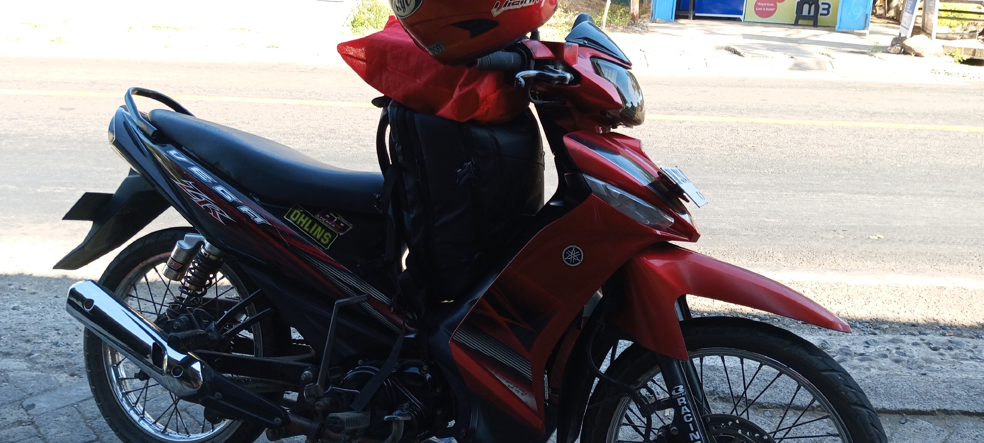 A red Yamaha Vega motorcycle is parked on the side of a road in Pakkanna, Indonesia. The motorcycle is equipped with a black seat, a chrome exhaust pipe, and a red helmet and bag are placed on top of the seat. The motorcycle has "OHLINS" and "YAMAHA" decals on its body. The road is paved with asphalt and bordered by a stone pathway. In the background, there are buildings and trees, suggesting an urban or suburban setting. The lighting indicates it is daytime with sunlight casting shadows. There are no people visible in the immediate vicinity of the motorcycle.