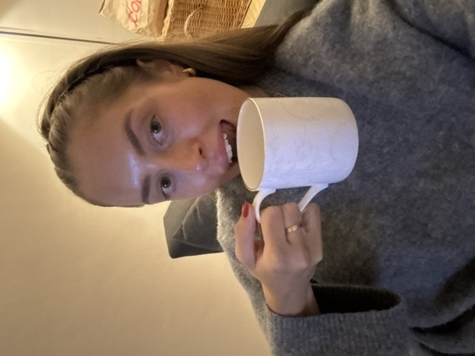 A woman in a gray sweater holds a white and gold mug to her mouth. She wears a black headband. Her left hand holds the mug handle, and a gold ring is visible on her finger. The background includes a wicker basket and a plain wall. The image is taken from a low angle.