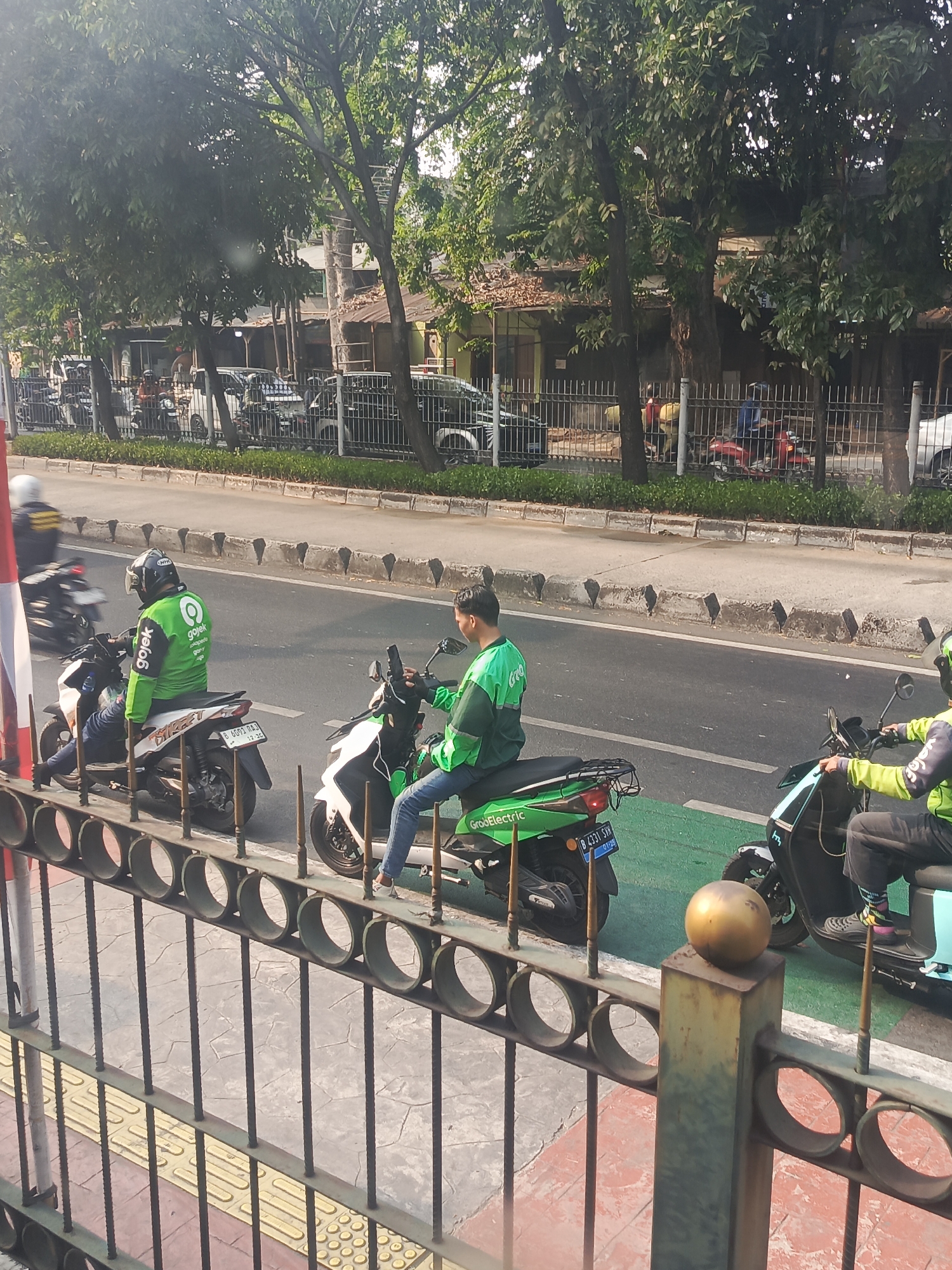 Two individuals on motorcycles are seen on a city street in East Jakarta, Indonesia. The person on the left, wearing a green jacket with a Gojek logo, is positioned on a white motorcycle. The person on the right, also wearing a green jacket, is on a green electric motorcycle labeled "GrabElectric." Both individuals are wearing helmets. The motorcycles are on a road with a green lane marking. In the background, trees line the street, and buildings are visible. A metal fence with circular openings is in the foreground, obscuring part of the view of the street. The scene appears to be captured during daylight hours.