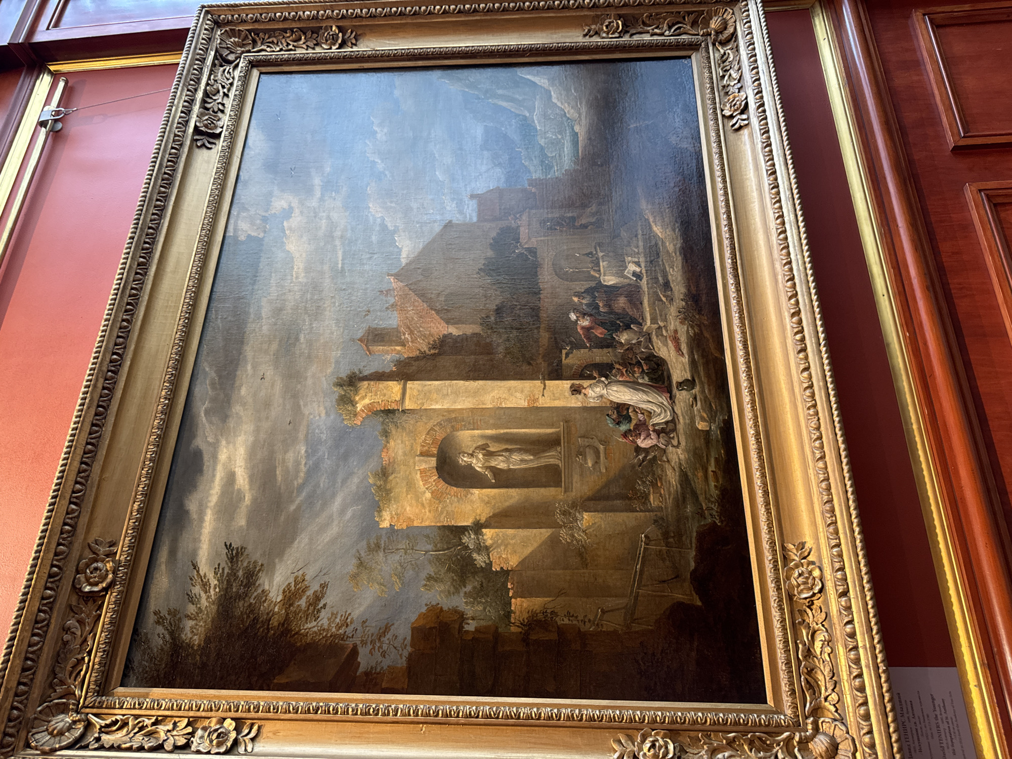 This is a painting of a historical or religious scene set in classical ruins under a dramatic sky. The central focus is a group of figures gathered around what appears to be a statue within an arched niche in a crumbling stone structure. Some figures are dressed in flowing robes, one in white, suggesting a religious or ceremonial event. To the left, a large, reddish building with a pitched roof dominates the middle ground, possibly a temple or dwelling, with a tower-like structure extending from it. The foreground is darker, with shadows and trees suggesting an outdoor setting. The sky is filled with dark, billowing clouds that create a sense of drama and possibly impending weather. The overall atmosphere is one of antiquity and perhaps melancholy, with the ruins evoking a sense of lost grandeur.
A small, partially visible label is attached to the lower right of the frame, with some text in what appears to be English and possibly another language, suggesting it might be an artwork in a museum or gallery. The wooden frame is ornate and gilded, with carved floral motifs. The lighting suggests it is daytime, but the heavy clouds contribute to a somber mood. The style of the painting, with its detailed rendering of architecture and figures, and the dramatic lighting, is reminiscent of Baroque or Neoclassical art.