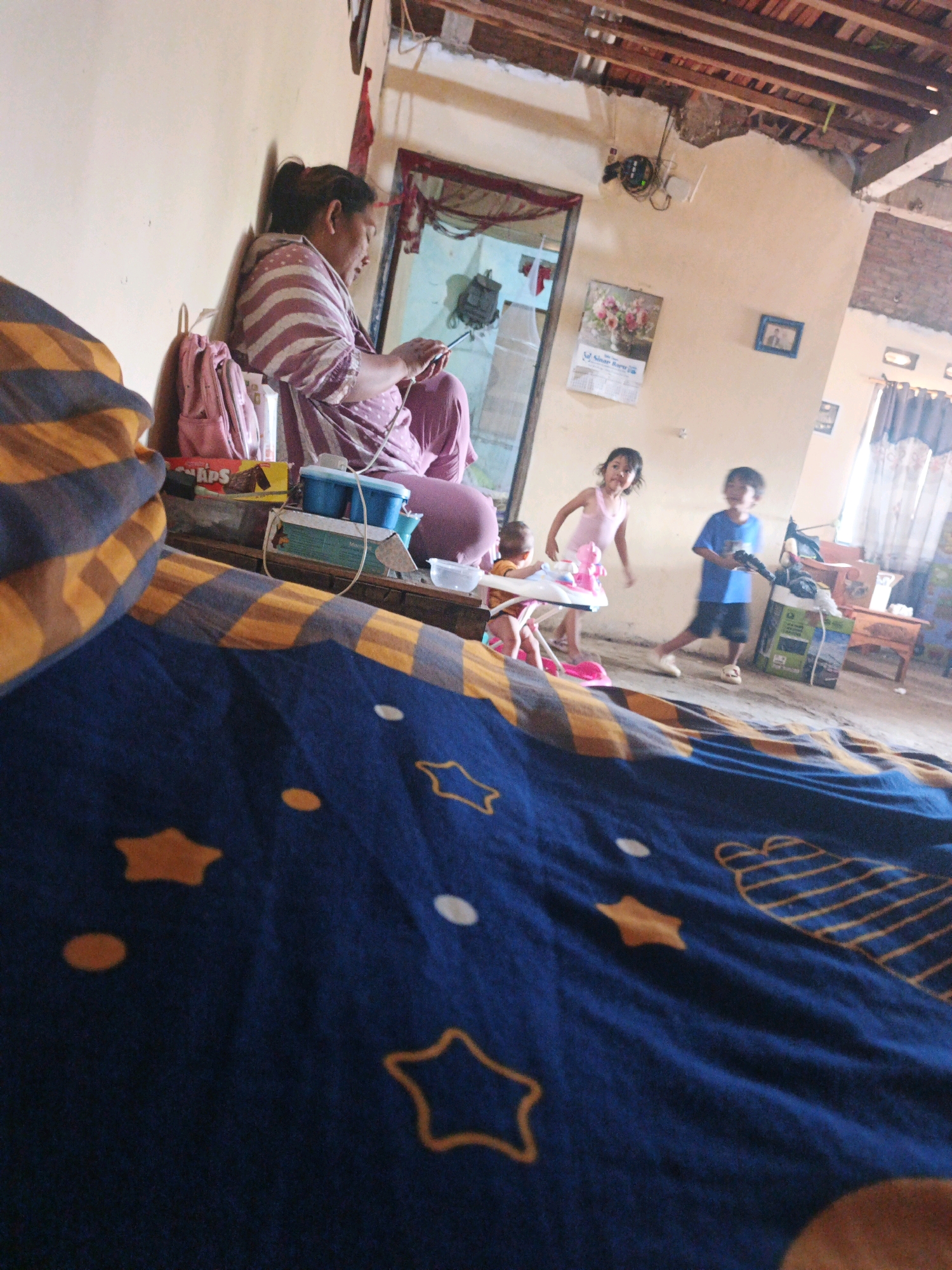 The image depicts an indoor scene, likely a living room or common area, within a home in Bumiharjo, Indonesia. In the foreground, a dark blue blanket with yellow stars and polka dots is spread out, taking up a significant portion of the lower half of the frame.
In the middle ground, a woman is seated on a raised platform or bed, leaning against a light-colored wall. She is wearing a purple and white striped shirt and purple pants. Her hair is tied back, and she is looking down at a smartphone she is holding in her hands. Next to her are a pink backpack, a box of snacks, and several blue plastic containers.
Further in the background, two children are visible. A young girl in a pink top is standing and appears to be playing or dancing. A young boy in a blue shirt and dark shorts is also standing. To the left of the children, a baby is seated in a pink and white baby walker. The environment appears to be simple and lived-in, with a concrete floor and unfinished ceiling elements. A calendar with a floral image hangs on the wall, along with a small framed picture. The overall impression is of a casual, everyday moment in a family home.