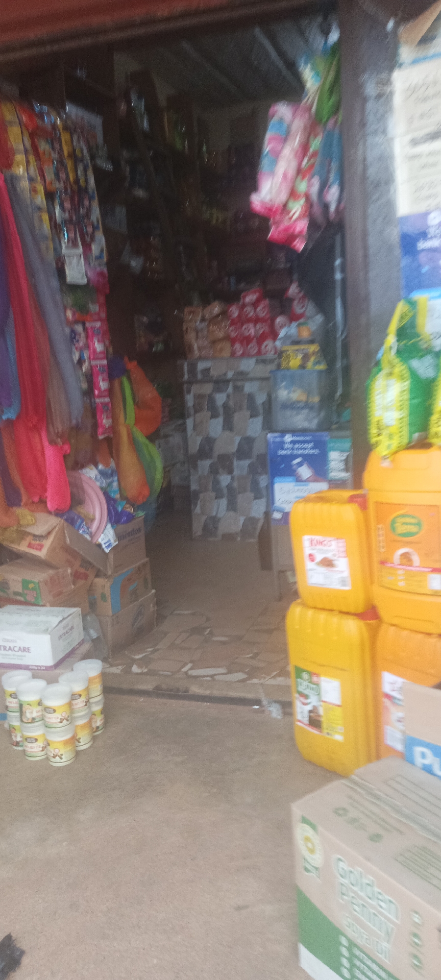 This image shows the entrance of a small shop located in Mangu Town, Nigeria. The shop is stocked with various goods, including foodstuffs, household items, and clothing. Some of the specific products visible include: vegetable oil, cooking ingredients, packaged foods, and draped fabrics. The entrance area is cluttered with boxes and items displayed for sale, and the overall atmosphere appears to be one of a busy, local retail environment.