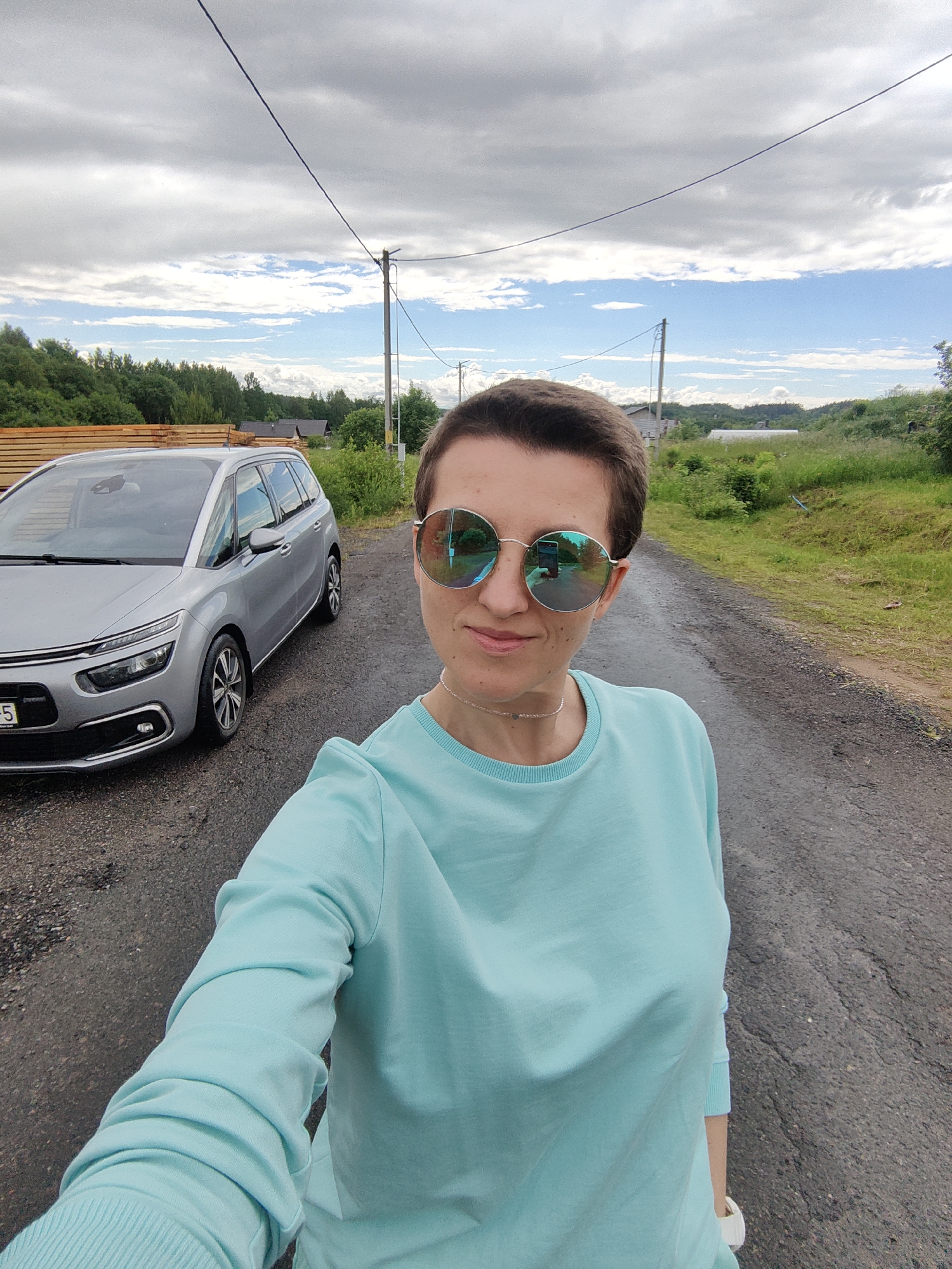 A woman is taking a selfie outdoors, standing on an asphalt road next to a silver minivan. She has short brown hair and is wearing round, reflective sunglasses and a light blue long-sleeved sweatshirt. Her necklace is visible around her neck. The minivan is parked on the left side of the road. In the background, there are trees, a cloudy sky, and some buildings or structures. The location is identified as Гаранскі сельскі Савет, Belarus.