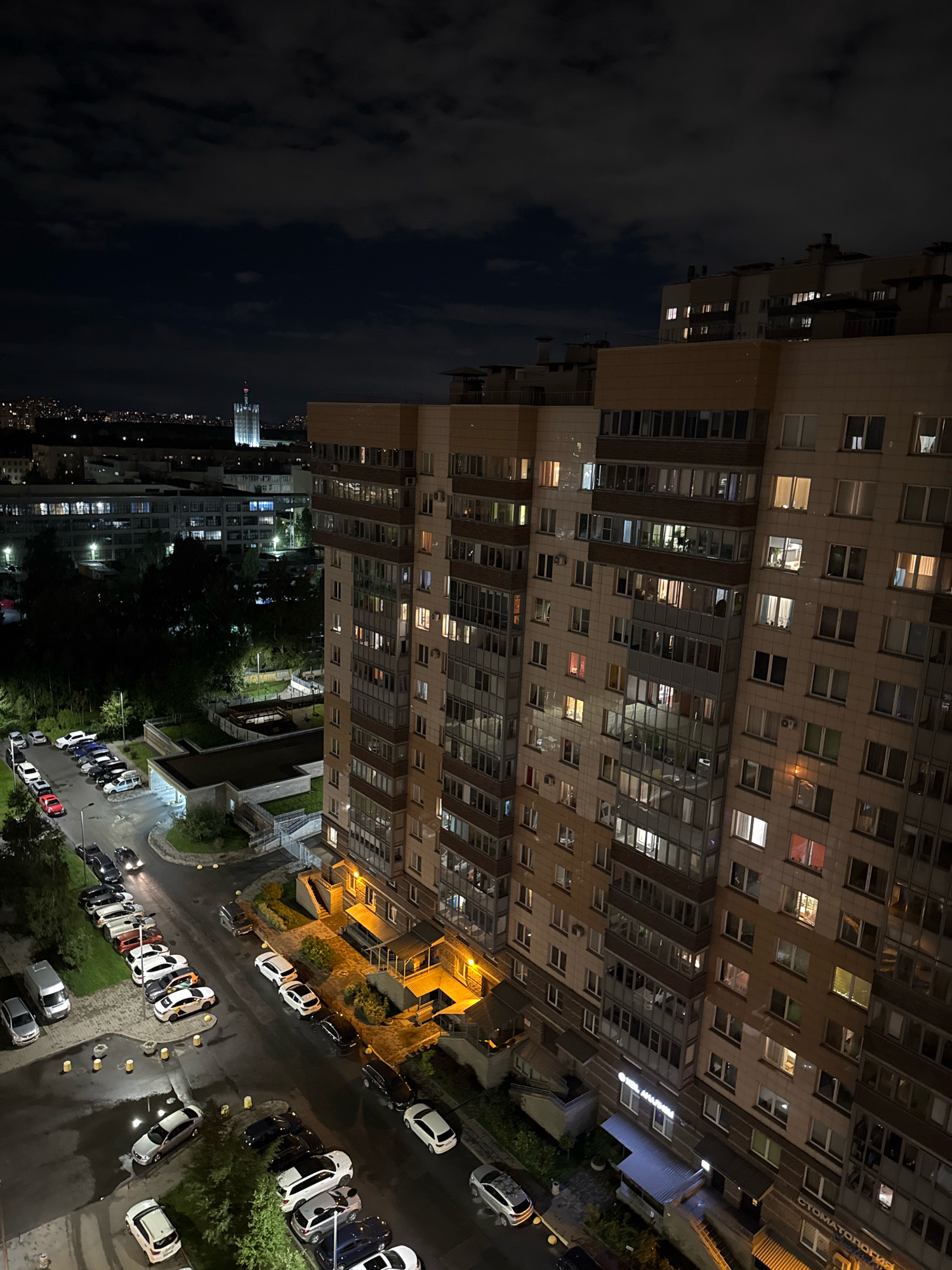 A night-time, high-angle aerial view captures an urban residential area in Shcheglovo, Russia. The scene is dominated by a large, multi-story apartment building with numerous illuminated windows. In the background, other buildings and a distant, tall, illuminated structure are visible, set against a dark, cloudy sky. Below the apartment building, a street is lined with parked cars of various models and colors. Some vehicles have their headlights on. Greenery, including trees and landscaped areas, surrounds the buildings and parking lots. The overall impression is one of a quiet, lit-up urban environment at night.