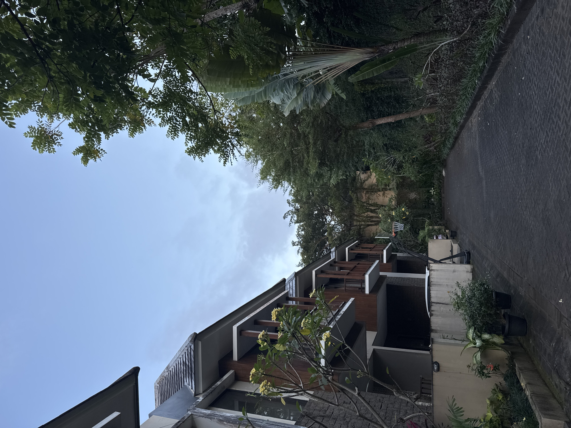 The image captures an exterior view of a modern residential building in Kuta Selatan, Indonesia. The multi-story structure has dark wood accents, gray concrete balconies, and a sloping roofline. A flowering tree with yellow blossoms stands in front of the building. Overhead, foliage from various trees frames the top of the image, creating a canopy effect against a partially cloudy sky. A paved walkway with a herringbone pattern runs alongside the building. Potted plants and greenery are visible along the base of the building's exterior.