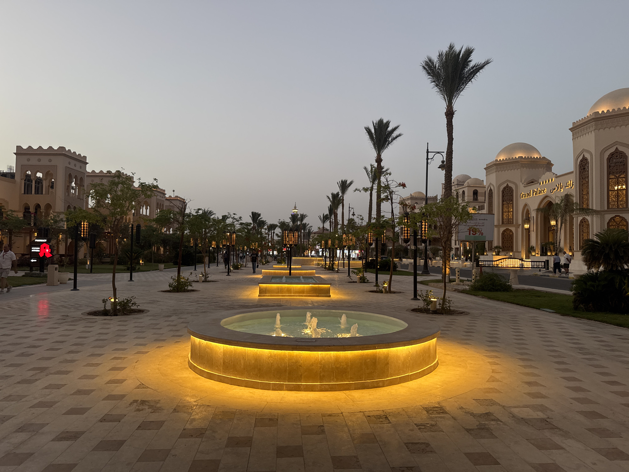 The image depicts a street scene at dusk in what appears to be a plaza or pedestrian area in a city. The scene features several round, lit fountains with water jets, positioned linearly along the central part of the street. Palm trees and street lamps are also aligned along the street, creating a sense of symmetry. On the right, a large building with ornate architectural details is visible, possibly a hotel or palace with "Grand Palace" signage. On the left, there are smaller buildings with distinct architecture, with some having arched windows. People are present, some walking in the distance. The paving stones create a pattern on the ground. The scene is generally well-lit, with a warm glow from the fountains and building lights contrasting with the fading daylight.