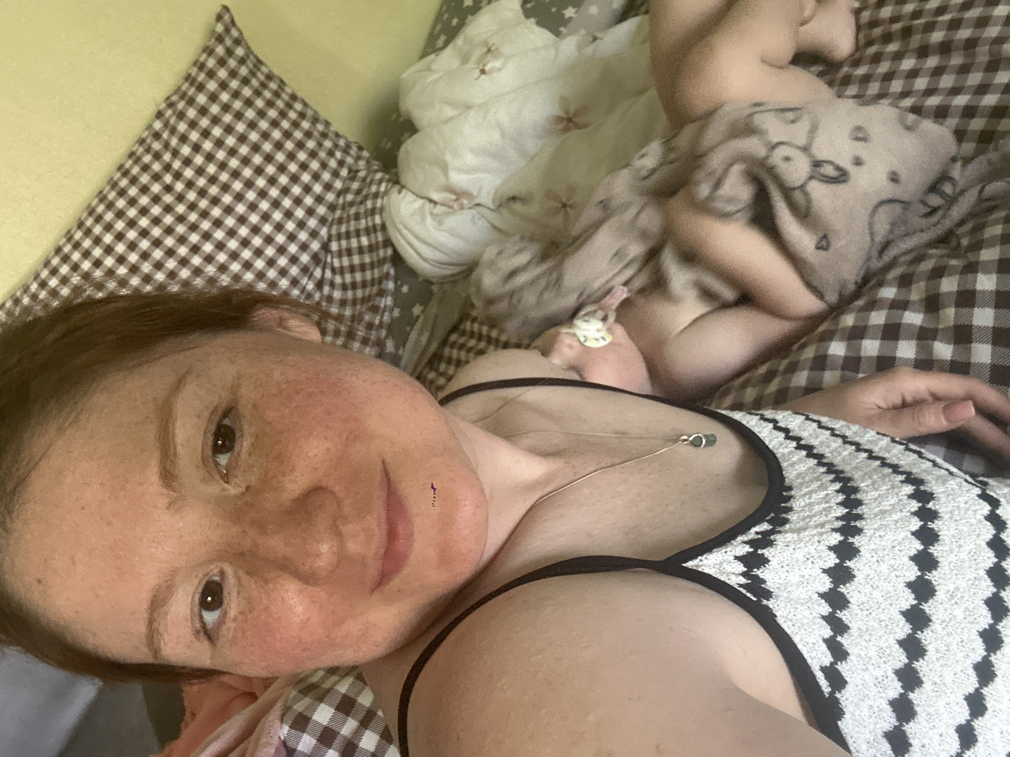 A selfie shows a woman in a white and black patterned top lying on a bed with brown and white checkered linens and a brown and white checkered pillow. A baby lies above her, partly covered by a blanket with a cartoon rabbit. A white blanket with floral patterns is also visible. The image was likely taken indoors, and there is no evidence to suggest a location in the city of Nizhny Novgorod, Russia.