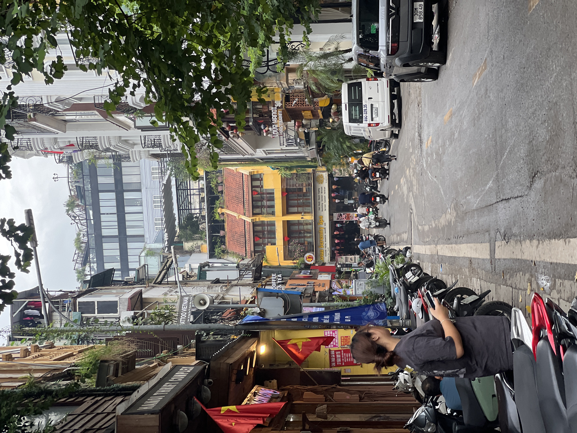 The image depicts a street scene in Hanoi, Vietnam, characterized by narrow buildings with ornate balconies, and a bustling street filled with motorbikes and a few cars. On the left side of the street, a woman stands looking at her phone, facing a row of parked motorbikes. Shops line the street, indicated by signs such as "HANOI THRIFT COMPLEX" and "TATTOO VIETNAM". Flags, including Vietnamese flags, are visible. Further down the street, a yellow building with orange-tiled roofs stands out. Several motorbikes are in motion on the road, along with a white van and a grey SUV. The sky is overcast.
