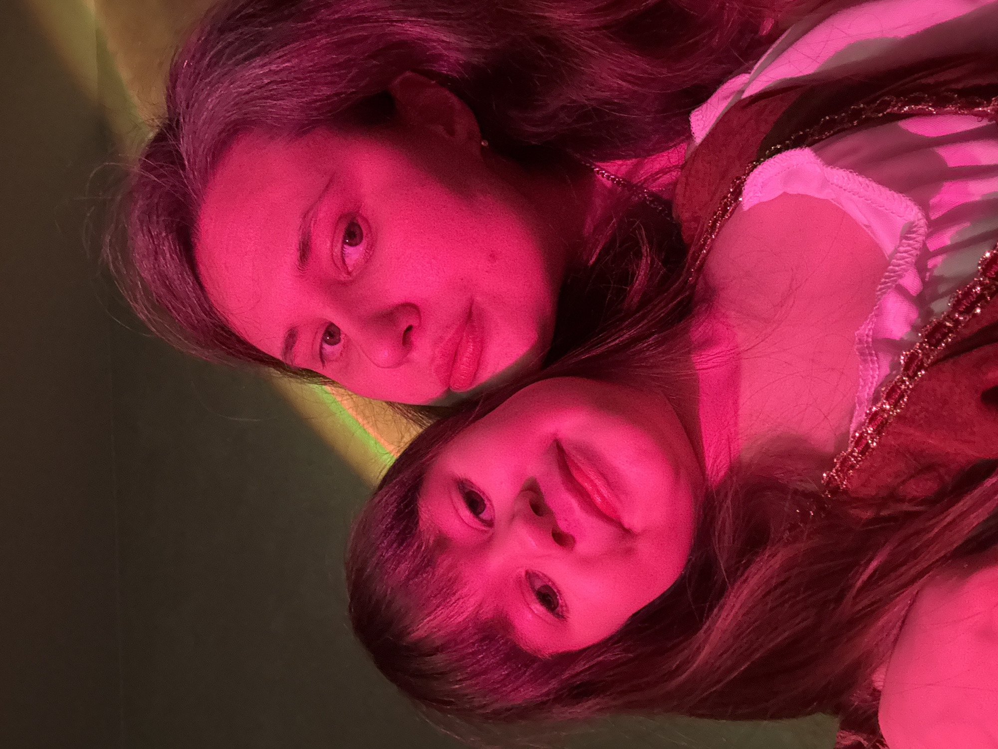 Two people, an adult and a child, are close together, possibly embracing. The adult is positioned above and slightly to the left of the child. Both are under strong, unnatural pink lighting. The background is a muted green. Details are difficult to discern due to the lighting. No information in the image relates to the city of Chaikovsky, Russia.