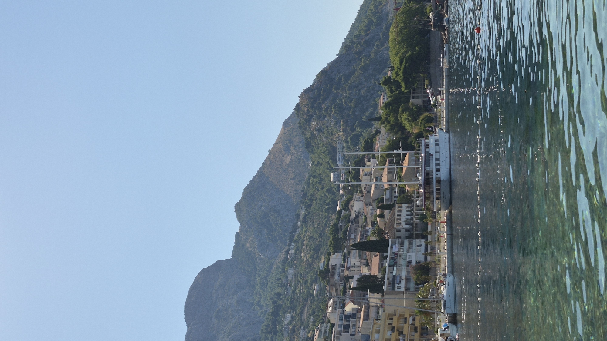 A vertical, eye-level, full shot captures a sun-drenched coastal town nestled against a verdant, mountainous backdrop under a clear, pale blue sky. In the foreground, the crystal-clear, turquoise water is disturbed by gentle ripples, with some white foam scattered across its surface. A light-colored buoy floats in the distance.
Along the shore, a white yacht with multiple decks and masts is moored. The word "VOYAGE" is visible in black lettering on its hull. Several other smaller boats are also docked. The town features a cluster of buildings with beige and white facades, some with red-tiled roofs, cascading down the hillside. Pine trees and other green foliage are interspersed throughout the urban landscape and the surrounding slopes. The mountains rise steeply behind the town, their rugged, rocky surfaces partially covered in dense, green vegetation. The overall impression is one of a peaceful, sunny day in a picturesque seaside location.