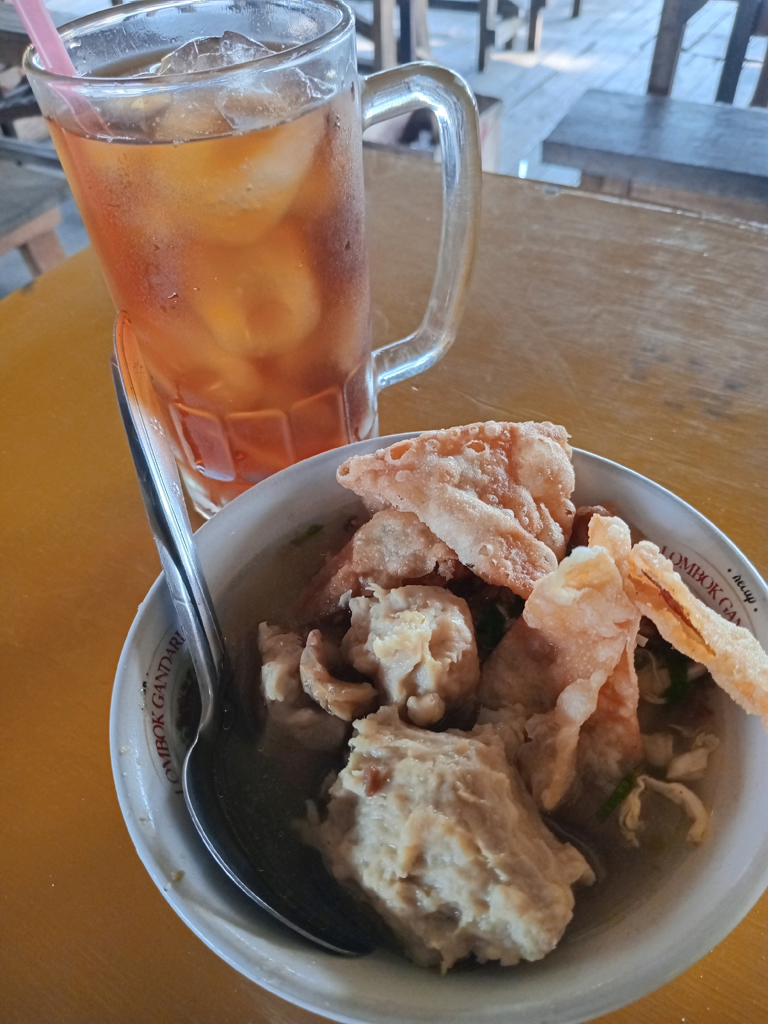 A tall glass of iced tea with a pink straw sits to the left of a white bowl of meatballs and broth. The glass is filled with amber liquid and ice cubes, and condensation is visible on the exterior. The bowl contains several large meatballs, some pieces of fried wonton or cracker, and a clear broth with what appear to be chopped green onions. A metal spoon rests in the bowl, angled towards the viewer. The bowl has text printed around its rim, which appears to read "LOMBOK GANDAR..." The items are placed on a wooden table. The background shows out-of-focus wooden furniture, possibly tables and chairs, suggesting an outdoor or rustic indoor seating area. The image is taken from a slightly elevated angle, looking down at the food and drink. There are no people visible in the frame.