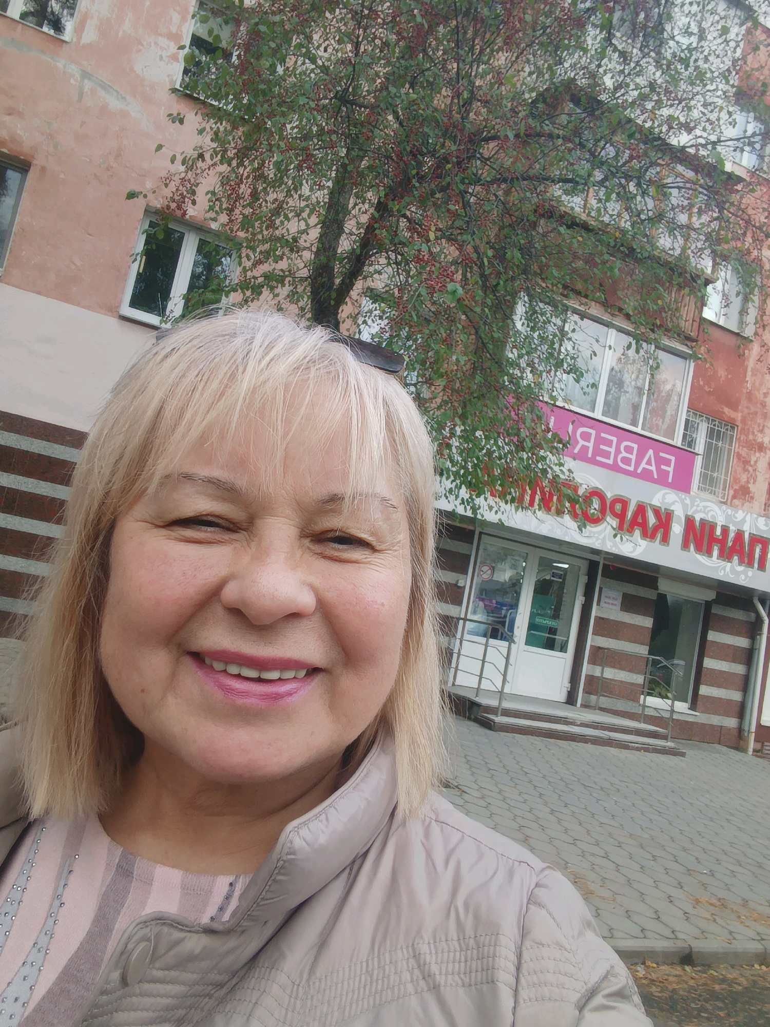 A middle-aged woman with blonde hair and a light-colored jacket is taking a selfie in front of a building in Asbest, Russia. The building has a pale pink facade and several windows. A tree with red berries is in front of the building, partially obscuring it. A sign on the building reads "FABERLIC" and "ПАН КАРОЛИ". The ground in front of the building is paved with grey interlocking stones. The woman is smiling and her makeup includes pink lipstick. The overall lighting suggests it is daytime.