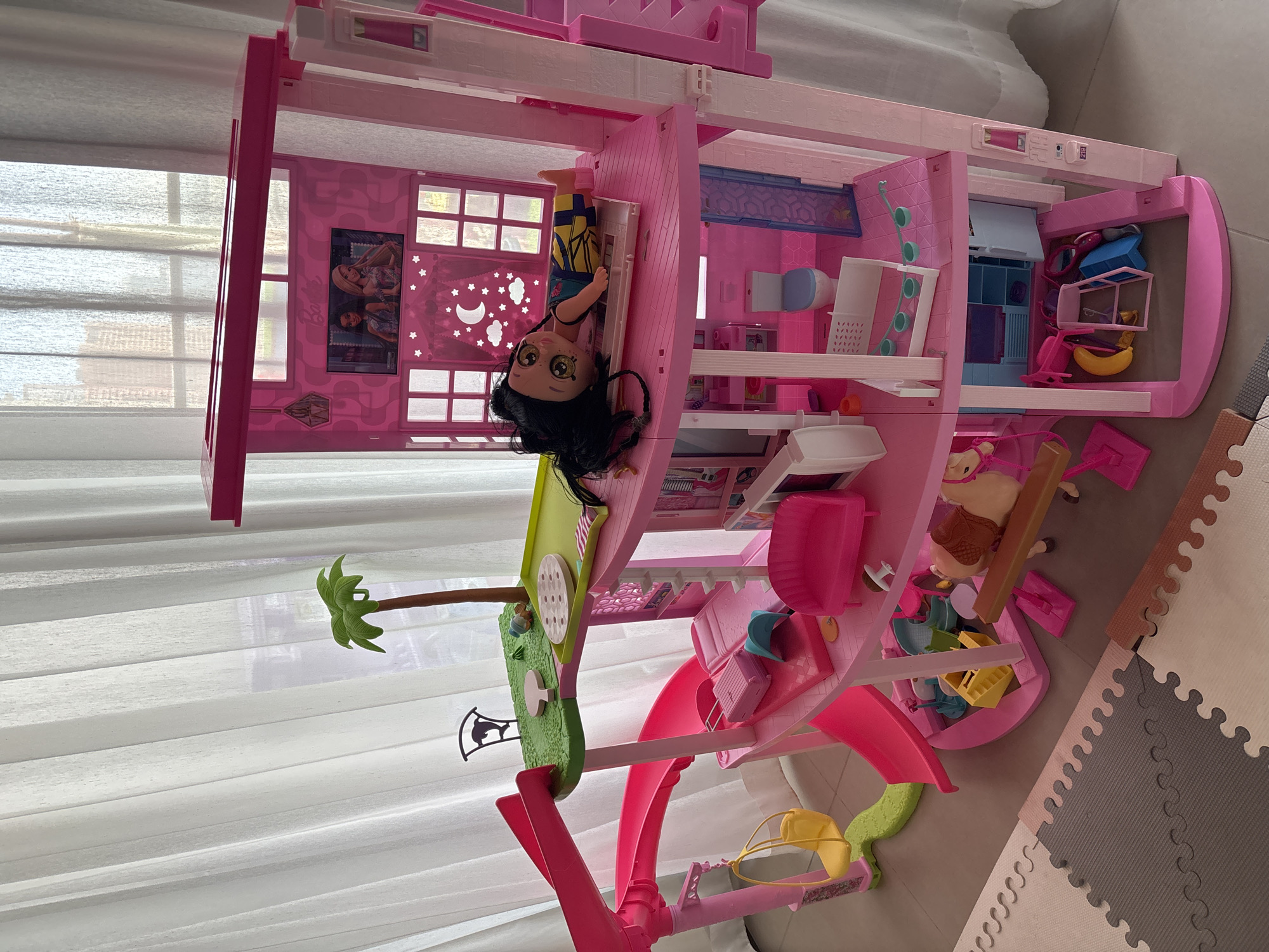 A large, pink plastic Barbie Dreamhouse is the central focus of the image, positioned indoors in front of sheer white curtains. The dollhouse features multiple levels, balconies, slides, and various accessories. On the uppermost level, a screen displays an image of two women, likely Barbie and a friend. A doll with dark hair is lying on a pink surface on this level. Below, other levels are furnished with a small bed, a pink sofa, a toilet, and various miniature items like a suitcase, a tent, a horse with a saddle, and a yellow swing attached to a slide. The dollhouse is set on a multi-colored foam floor mat, suggesting a child's play area. The background visible through the curtains hints at an urban environment with tall buildings, consistent with the city of Dubai, United Arab Emirates. The lighting suggests it is daytime.
