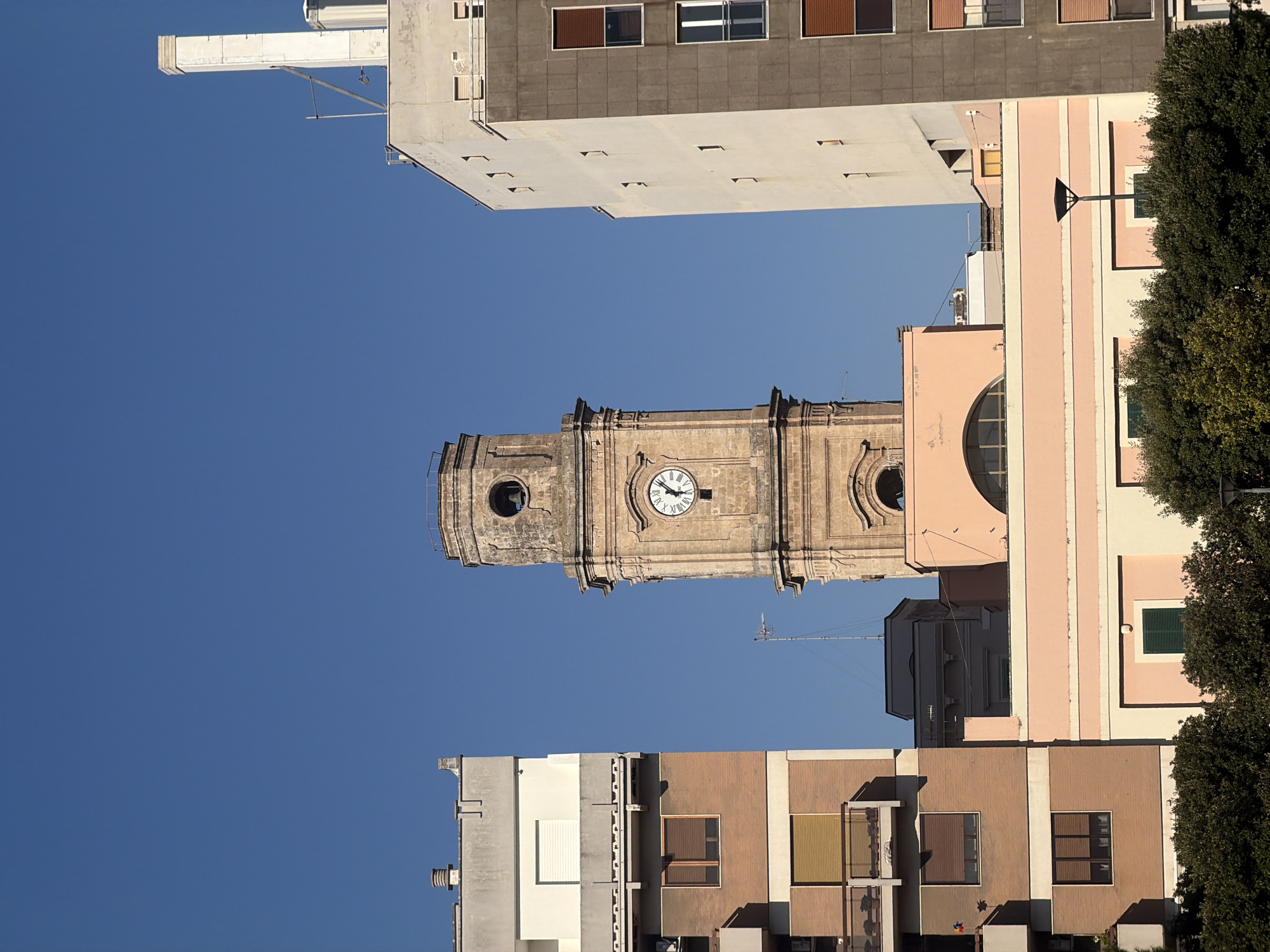 The image shows a street view in Monopoli, Italy, focusing on a historic clock tower situated between two modern buildings. The clock tower, made of weathered stone, has a circular opening with a bell above the clock face. The clock face itself displays Roman numerals and hands indicating approximately 11:05. To the left, a residential building with a brick facade and balconies is visible. To the right, a taller, modern building with a gray and white exterior stands prominently. The foreground is dominated by lush green trees and bushes, with a lamppost visible on the right side. The sky is clear and blue, indicating a sunny day.