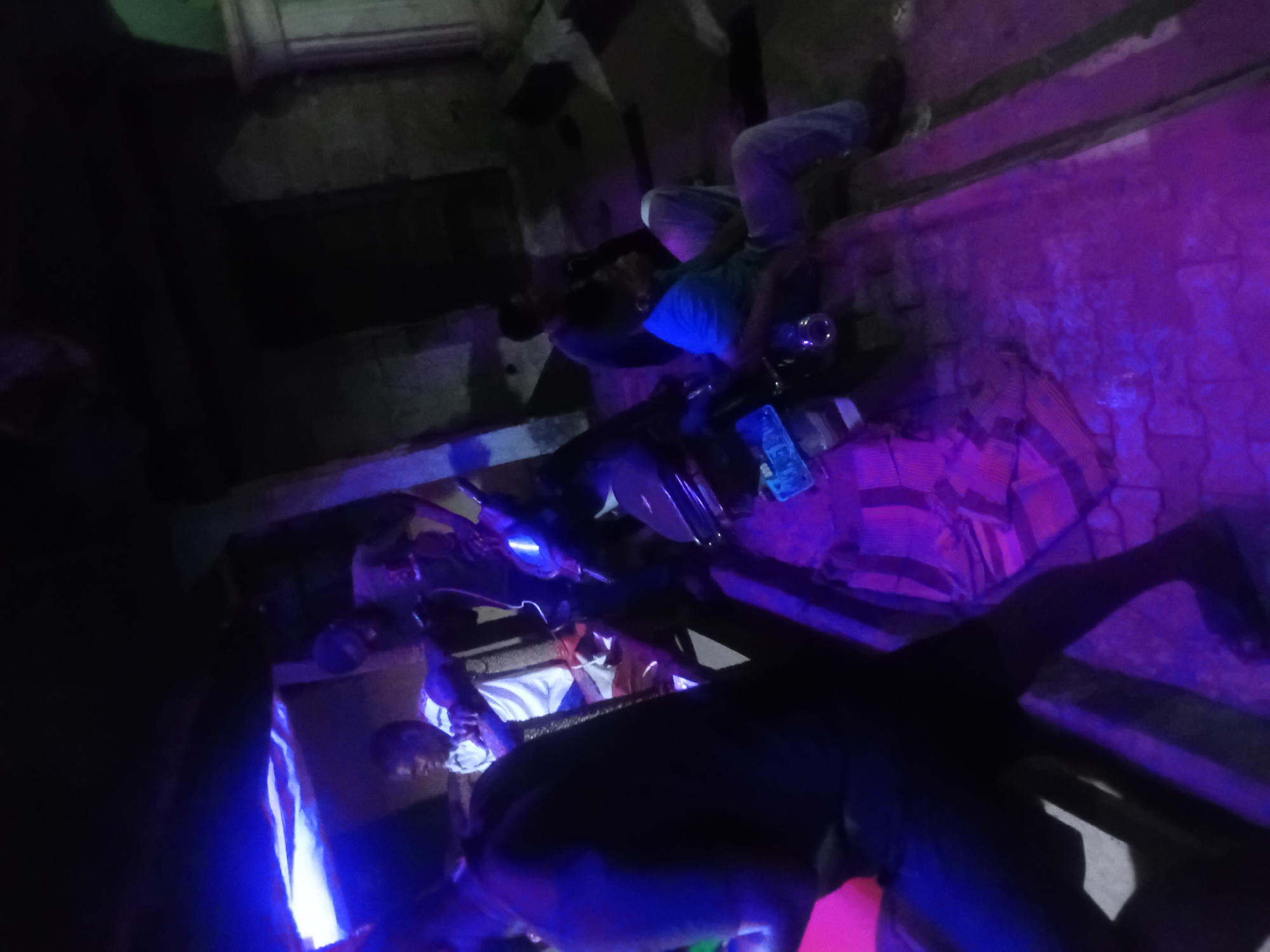 The image shows a group of individuals in an indoor environment, likely a staircase or narrow passage. The scene is illuminated with blue and purple light, possibly from artificial sources. Several people are visible, some lying or sitting on the steps. One individual is wearing a blue shirt and light-colored pants. Other people are dressed in dark clothing. A motorcycle is also visible in the frame. Several water bottles are visible in the center of the image. It is difficult to determine the exact nature of their activities due to the darkness and the angle of the shot. The setting appears to be a concrete structure with uneven steps.