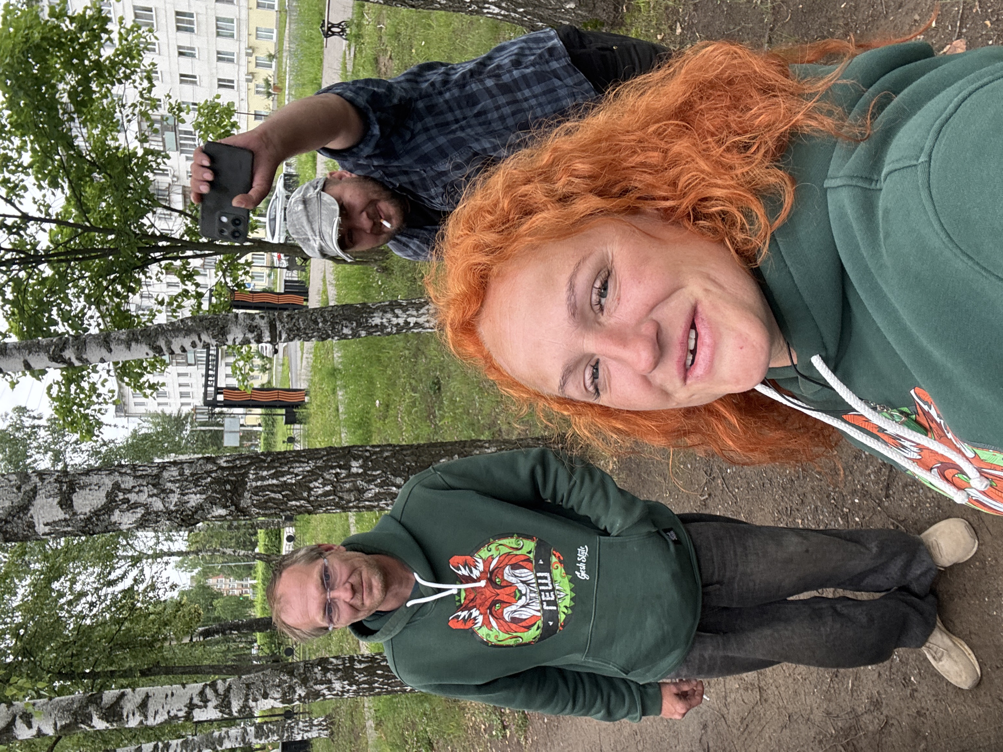 In Chaikovsky, Russia, three individuals are captured in an outdoor photograph. The central figure in the foreground is a woman with vibrant red, curly hair and fair skin, wearing a green hooded sweatshirt. To her left, a man with short, light brown hair, wearing glasses and a similar green hooded sweatshirt, stands looking towards the camera. In the background, a man wearing a plaid shirt and a cap is holding up a black smartphone, seemingly taking a picture or recording. He also appears to be smoking. They are situated in a park-like setting with birch trees, grass, and a residential building visible in the distance. A decorative element resembling the Order of Saint George ribbon is also present. The ground appears to be a mixture of dirt and gravel.