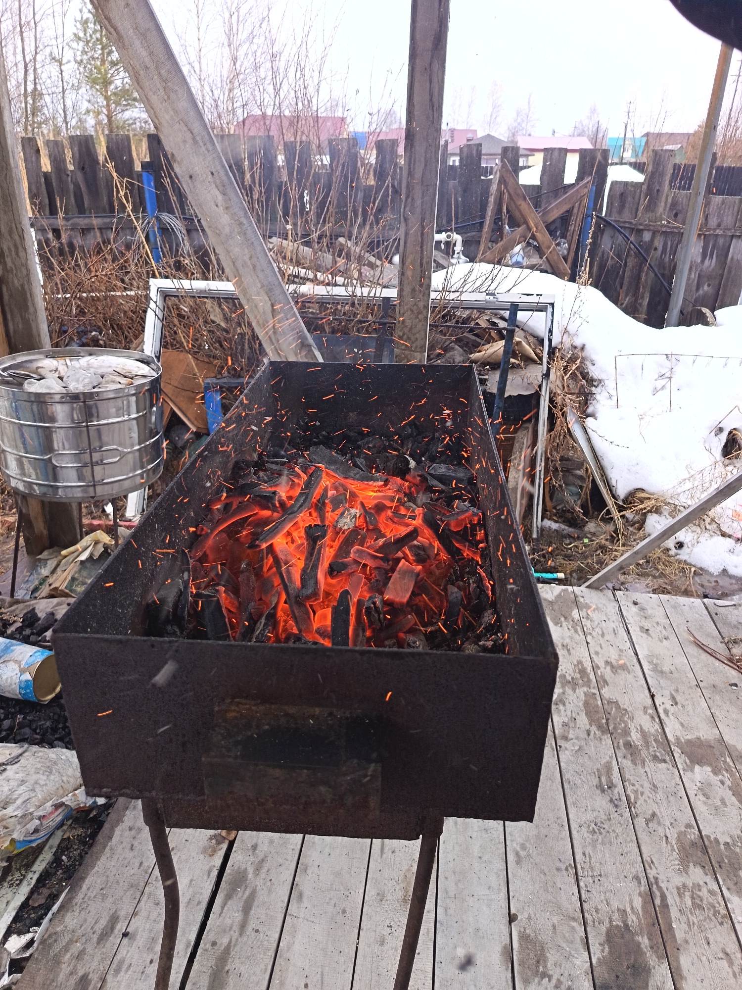 The image shows a metal brazier filled with burning embers, likely charcoal, on a wooden platform. Sparks are visible rising from the fire. A metal container with a lid sits to the left of the brazier. In the background, there's a wooden fence and some bare bushes or small trees. Patches of snow are visible near the fence.
Given the reference to Nizhnevartovsk, Russia, it is possible this scene is situated in a backyard or a semi-urban area with a rustic, possibly temporary, structure. There are no visible people in the image. The brazier is the central subject, suggesting an outdoor cooking activity.