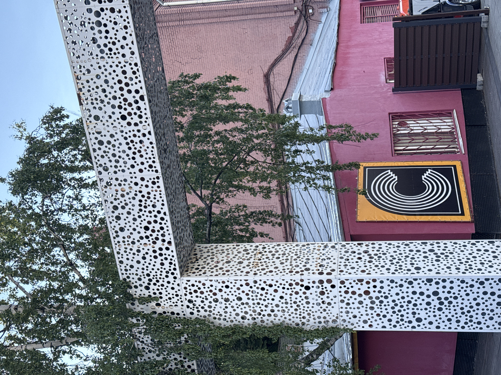 A street view in Moscow, Russia, shows a structure with a design of varying sized holes, adjacent to a pink brick building. A tree with green leaves is visible through the holes and adjacent to the building. A sign with concentric white semi-circles on a black background is attached to the pink building, below a window with bars. A covered balcony or deck is at the top of the building. The sky is light blue.