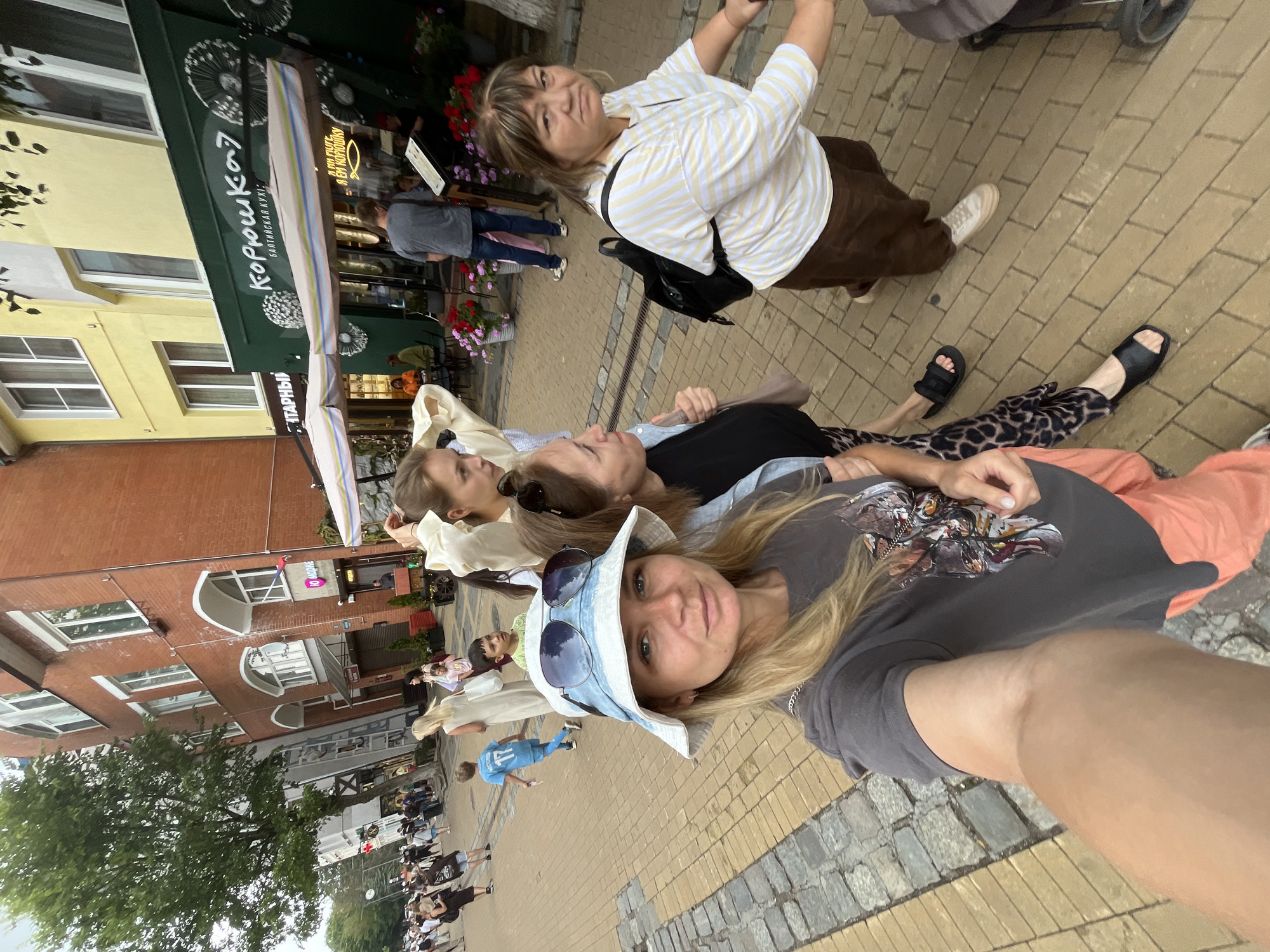 In Zelenogradsk, Russia, on a brick and cobblestone sidewalk, four individuals are captured in a street scene. In the foreground, a woman wearing a white bucket hat and a grey t-shirt with a graphic print is taking a selfie. She is accompanied by two other women; one in a black shirt with leopard-print leggings and black slides, and another in a light-colored striped shirt and brown pants. To the right, a stroller is partially visible, pushed by an unseen person. In the background, a street lined with brick and yellow buildings features outdoor seating for a restaurant with green awnings, bearing the Cyrillic sign "КОРОШКА?". Several people are visible walking on the sidewalk further down the street. The scene appears to be a daytime, casual outing.