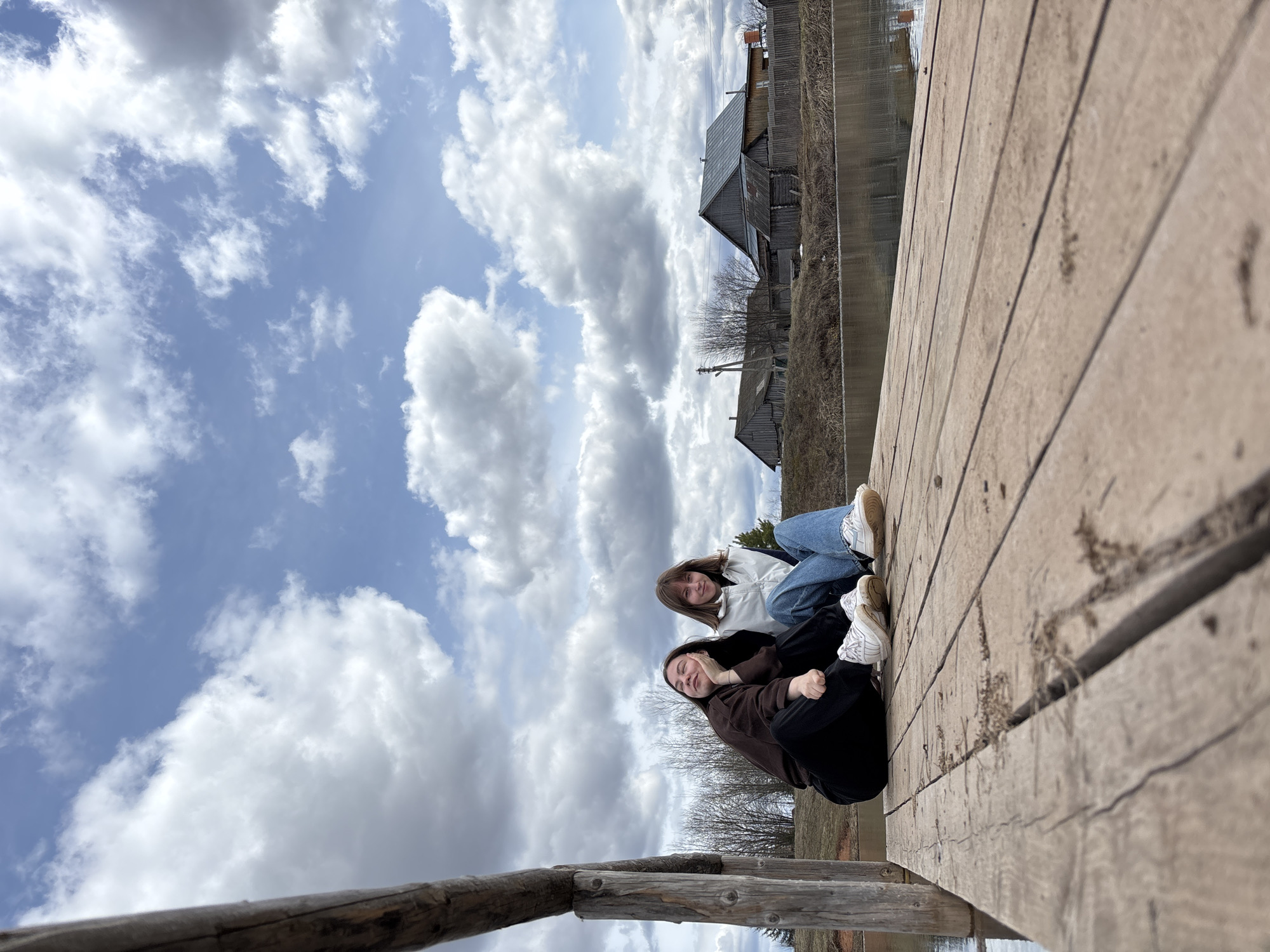 This photograph, set in Петропавловск, Russia, features two young women sitting casually on a rustic wooden pier. The pier, made of aged, textured wooden planks with visible grain and some dirt in the crevices, extends from the lower right of the frame towards the upper left, forming a prominent foreground element.
The two women are positioned towards the left side of the pier. The woman on the right wears a light-colored (possibly white or light grey) jacket, blue jeans, and white sneakers. She has medium-length brown hair and looks towards the camera with a soft smile. Beside her, the woman on the left is dressed in a dark brown top or jacket, black trousers, and white sneakers. She has longer, darker hair and rests her head on her hand, also looking at the camera with a relaxed and content expression. They appear to be posing for a casual photograph.
Beyond the wooden pier, a calm body of water, likely a river or lake, fills the midground, subtly reflecting the sky above. On the far bank, several traditional wooden houses with dark, sloped roofs are visible, nestled among bare trees. The absence of leaves on the trees suggests an early spring or late autumn season. The sky is a significant feature, characterized by large, fluffy white and grey clouds against patches of bright blue, indicating a partially cloudy but bright daytime. The overall scene conveys a sense of peaceful outdoor leisure in a scenic, rural or semi-rural environment. No text is visible in the image.