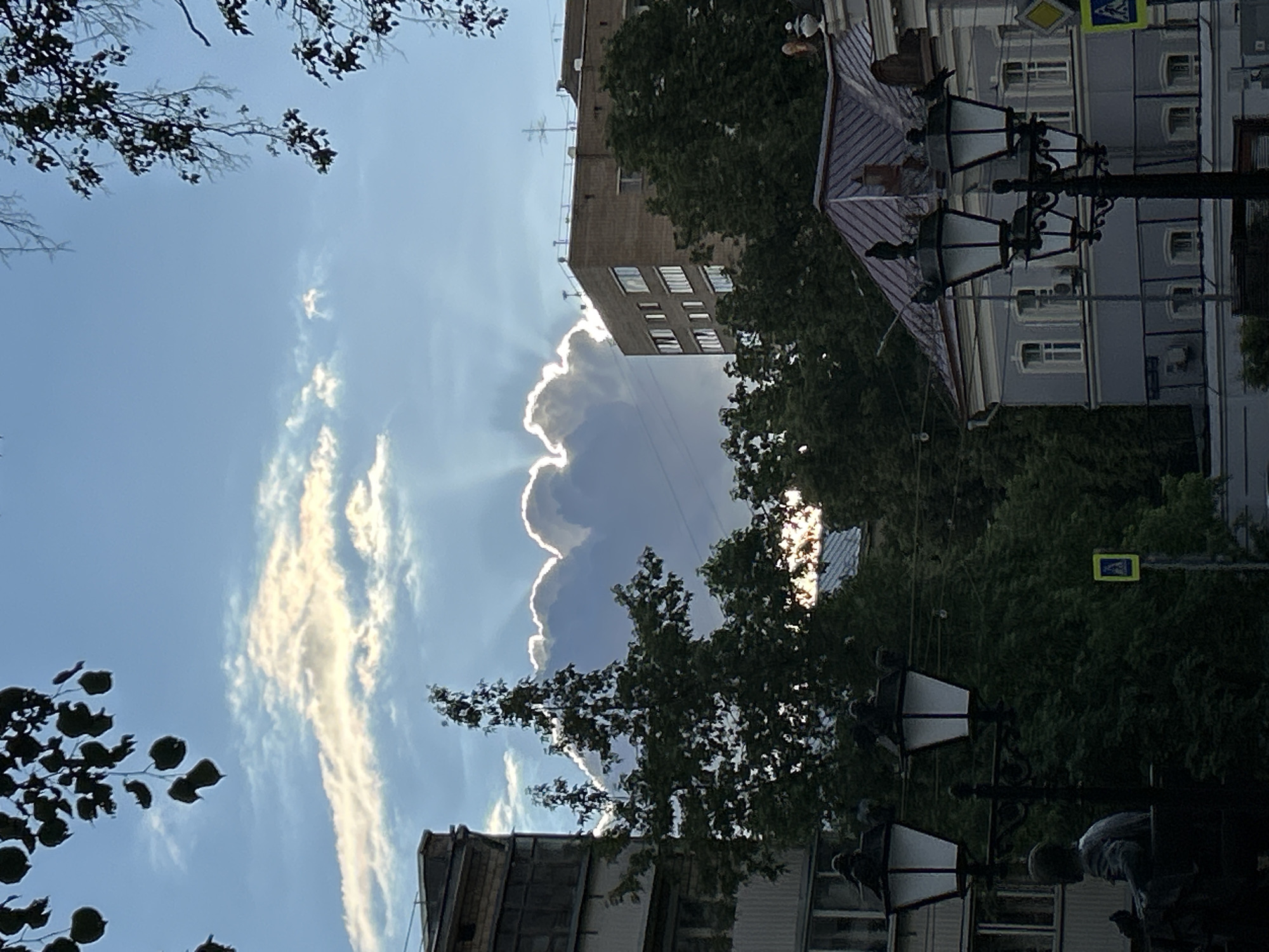 The image shows a view of a city street in Unknown, Unknown, looking upwards towards the sky. Buildings are visible on both sides of the street, partially obscured by trees. A large, dark cloud with bright, sunlit edges dominates the center of the frame. Streaky, elongated clouds are also present in the blue sky to the left. Street lamps with white shades are visible in the foreground and along the right side of the frame. A yellow and blue pedestrian crossing sign is also visible.