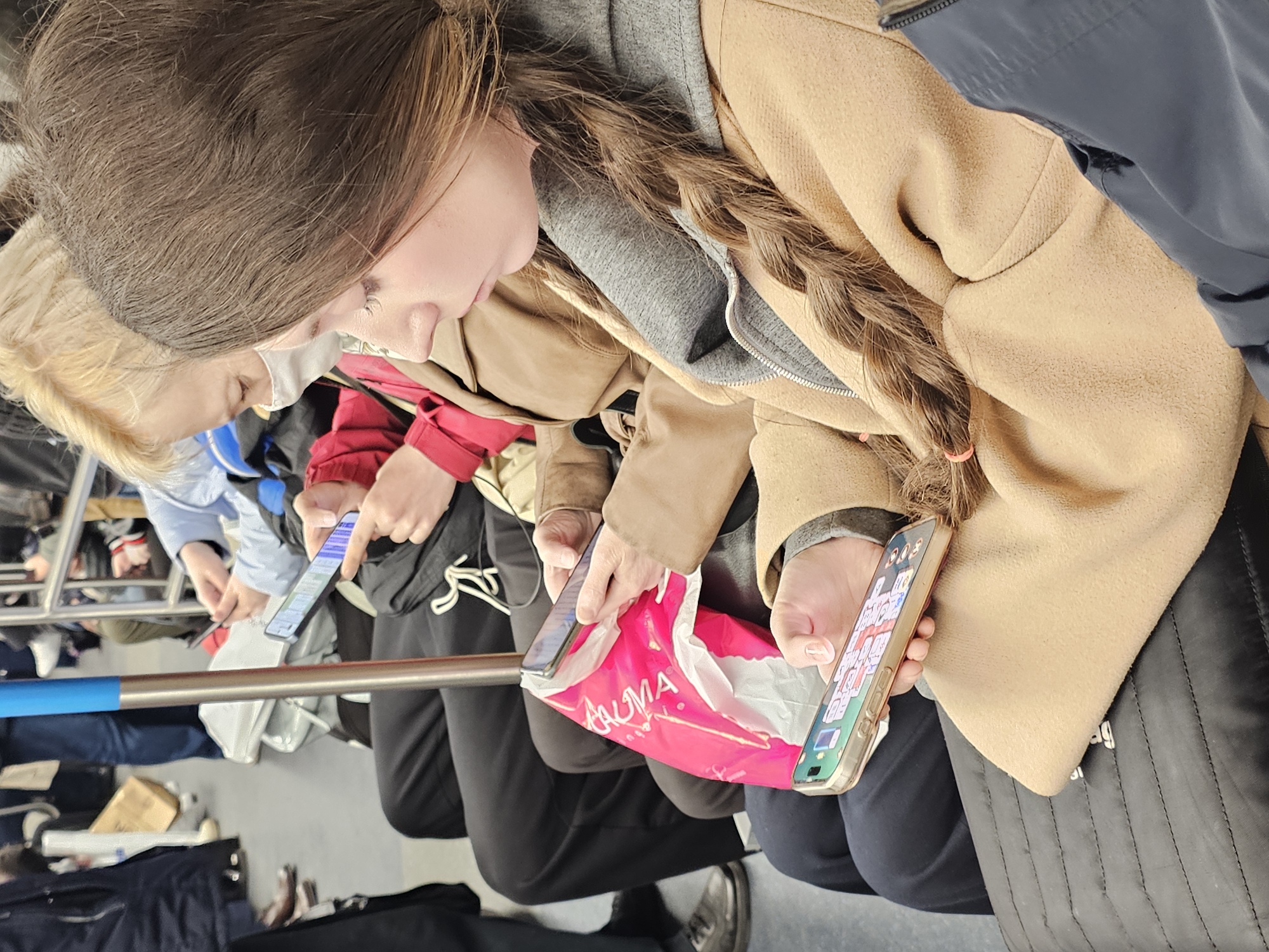 The image captures several people, likely on public transport or in a crowded waiting area, with a focus on their engagement with smartphones. In the foreground, a woman with long brown hair, styled in a braid, wears a tan coat over a grey hooded top. She is looking down, engrossed in her smartphone, which is held in her right hand and displays an interface with numerous small icons.
To her immediate left, another person with blonde hair is partially visible, also looking downwards, suggesting they are also using a device. Further back, more individuals are present, with at least two others clearly holding and operating their smartphones. One person, to the left of the main subject, holds a phone displaying what appears to be a messaging or social media application.
A bright pink bag is visible in the mid-ground, nestled between individuals, with the white text "TALMA" prominently displayed on it. A blue and silver metal railing extends horizontally across the frame, dividing the foreground from the background. The scene is indoors, suggesting daytime lighting. The subjects appear focused on their phones, conveying a sense of individual engagement within a shared public space.