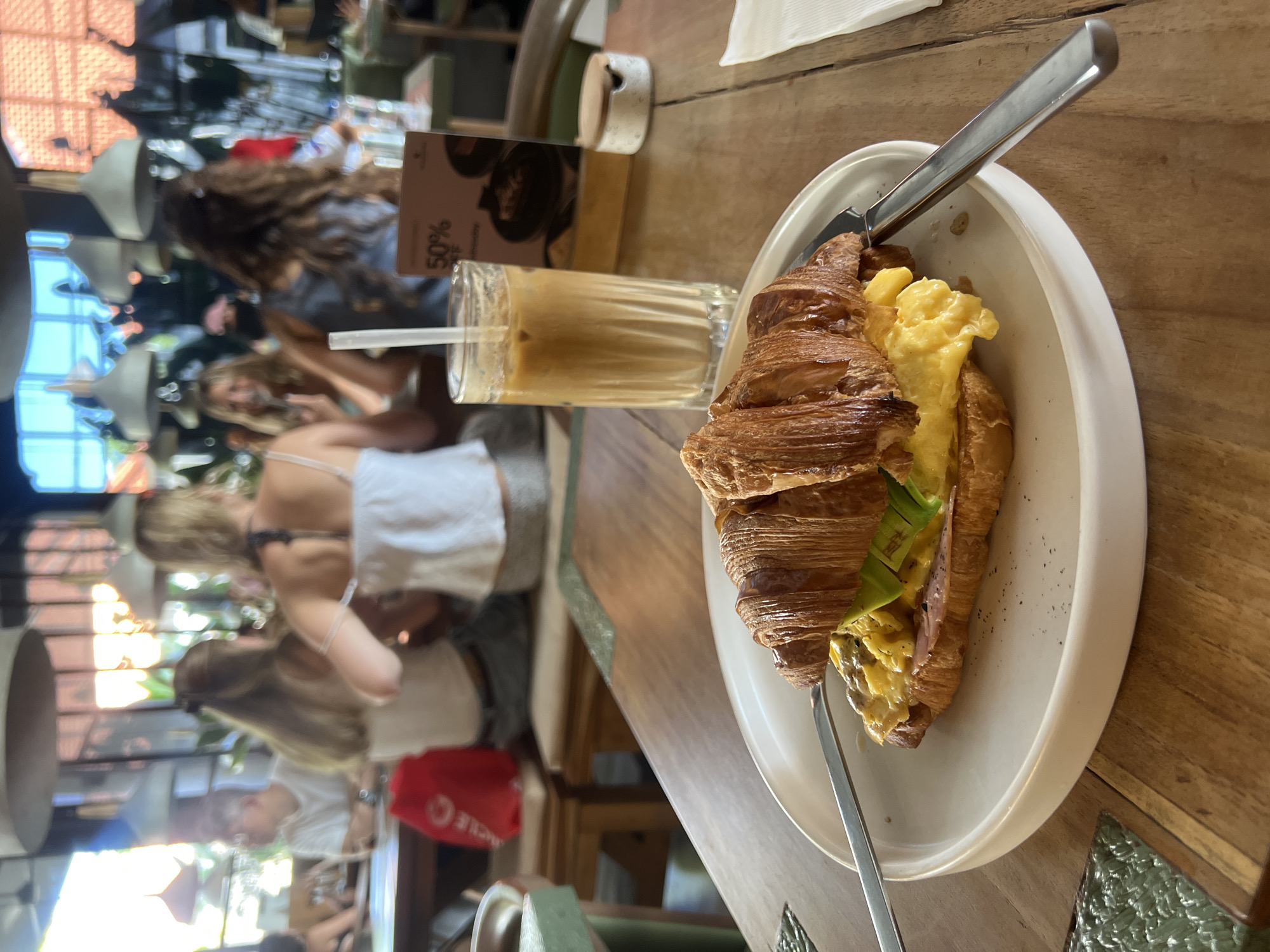 The image shows a table in a café in North Kuta, Indonesia. A croissant sandwich with scrambled eggs and avocado is on a white plate, accompanied by a latte in a glass with a straw and two knives. In the background, blurred figures of people are sitting at tables, with a window and hanging lights visible. The table is made of wood.
