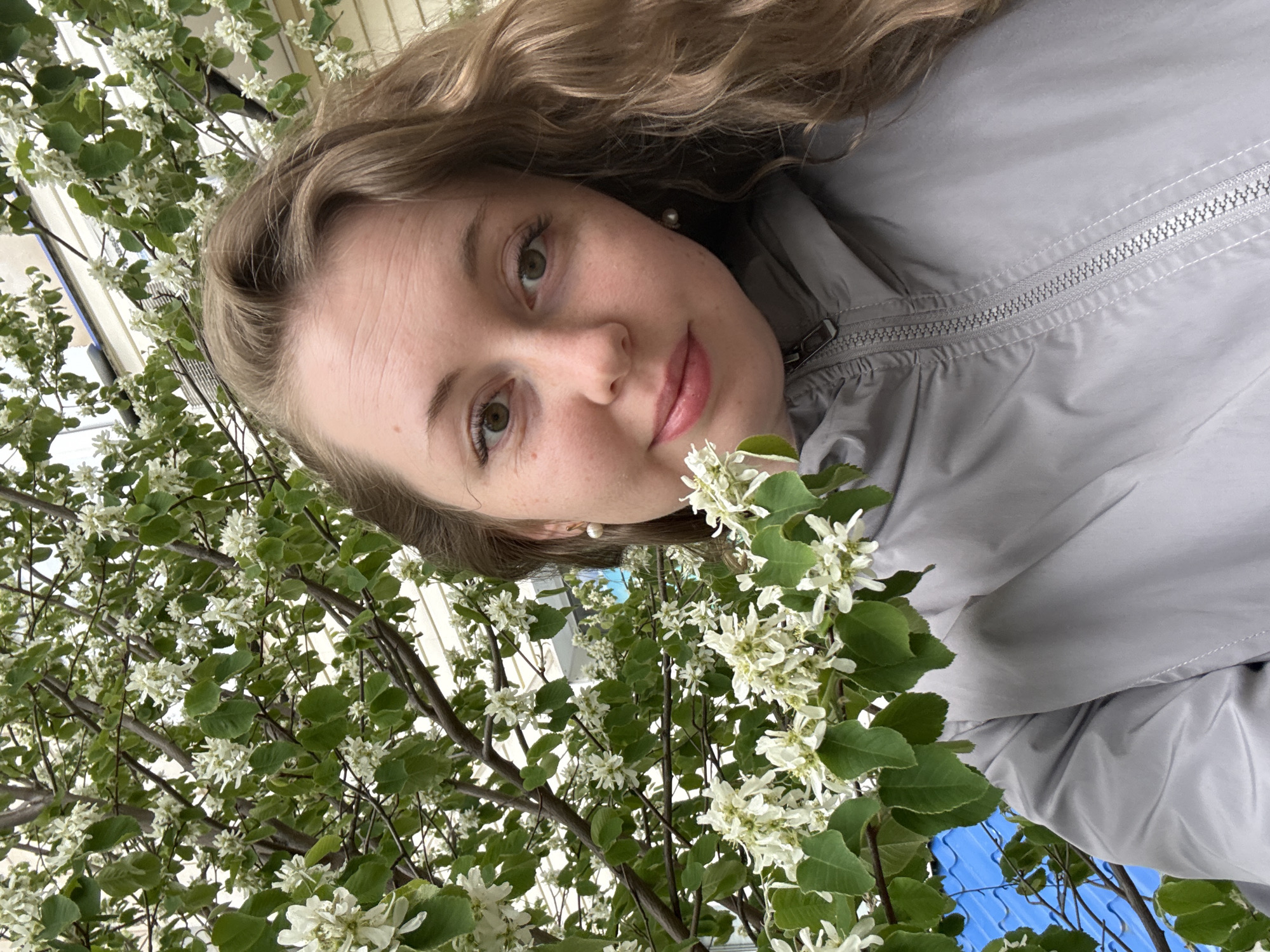 A fair-skinned woman with curly blonde hair stands near a flowering tree in Unknown, Unknown. She wears a gray jacket and white pearl earrings. The tree has green leaves and small white flowers. A building with white siding is visible in the background, along with a patch of blue sky.