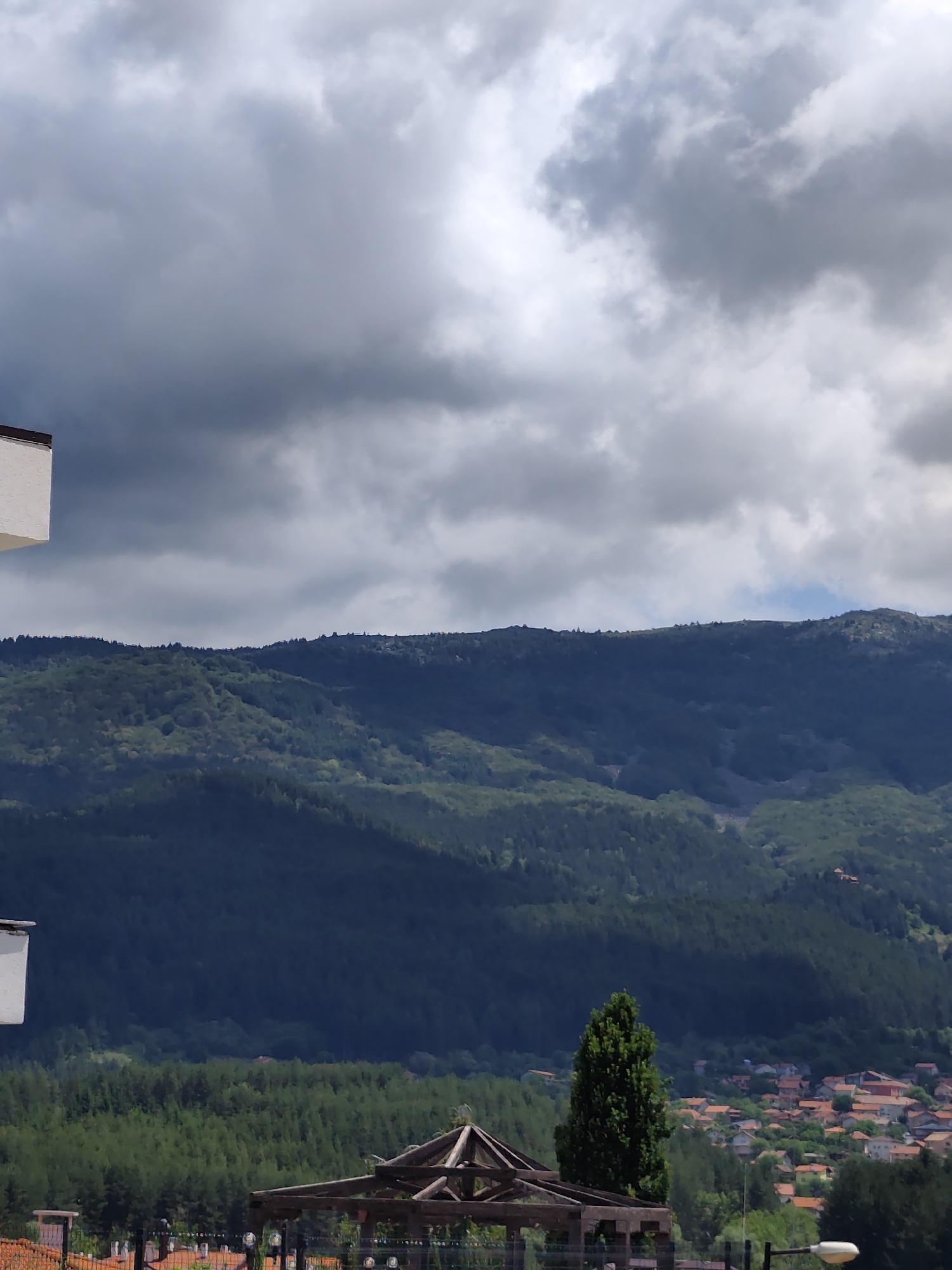 The image depicts a mountainous landscape with a village nestled in the valley, as seen from Kladnitsa, Bulgaria. The sky is filled with dramatic, dark clouds, suggesting an overcast or potentially stormy weather condition. In the foreground, a portion of a white building is visible on the left. Below the building and extending across the lower part of the image, a dense forest of green trees covers the slopes of the mountains. A small, isolated tree stands prominently in the mid-ground to the right of the center. The village, characterized by its terracotta-tiled roofs, is scattered across the lower slopes and valley floor. A partially constructed wooden gazebo or pergola structure is visible in the bottom center, with a black metal fence in front of it.