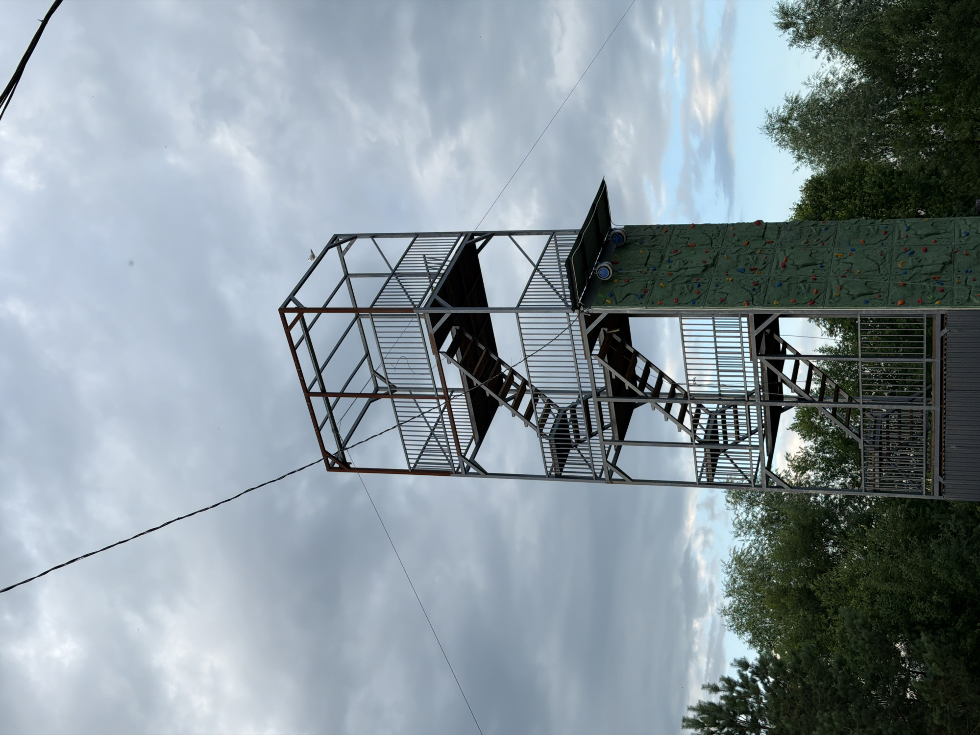 A tall, metallic climbing tower stands against a cloudy sky in Bol'shaya Ven'ya, Russia. The tower has multiple levels with metal stairs and railings. A green climbing wall with colorful holds is attached to one side of the tower. Trees surround the base of the tower. A thin wire extends diagonally across the upper portion of the frame.