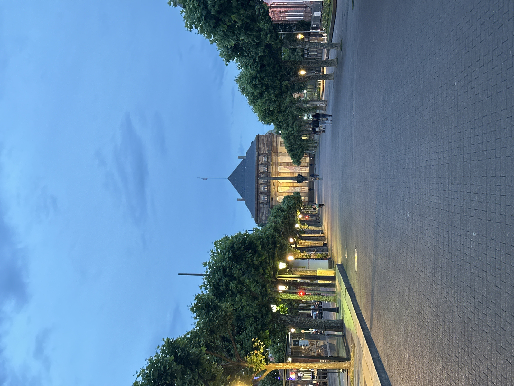 Captured at dusk or twilight, the image portrays a grand urban street or plaza in Strasbourg, France. The sky is a deep, tranquil blue, marked by subtle cloud formations, as artificial lights begin to illuminate the scene.
In the foreground, a wide, meticulously paved area stretches into the distance, composed of dark gray rectangular pavers, suggesting a substantial pedestrian thoroughfare or esplanade. This paved expanse is bordered on both sides by symmetrically planted rows of mature trees, featuring dense green foliage. Interspersed among the trees are numerous streetlights, casting a warm, inviting glow that contrasts with the cool blue of the twilight sky. A red traffic light can be faintly observed through the foliage on the right side.
Several small figures, appearing as people, are scattered in the mid-ground and background, walking along the paved area or near the structures. At the far end of this vista stands a prominent, classical-style building, characterized by its ornate stone facade and a distinctive dark, pyramid-shaped roof topped with a flagpole. The building's windows are softly lit, contributing to the evening ambiance.
The scene conveys a peaceful and calm atmosphere, with the visible activity limited to a few individuals strolling. The symmetrical arrangement of the tree-lined avenue and the interplay of natural and artificial light are notable details, enhancing the sense of urban grandeur. No specific text is clearly discernible or readable on any signs or structures within the image.