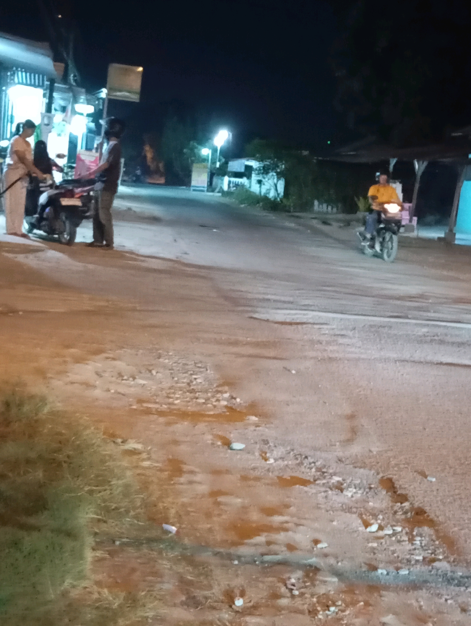 The image depicts a nighttime scene on a paved road, possibly in Leman Benawi, Indonesia, characterized by low-light conditions and some reflective surfaces on the ground. On the left side of the road, two individuals are near a motorcycle. One person, appearing to be a woman in light-colored clothing, is standing near the rear of the motorcycle. Another person, a man in darker clothing, stands beside her, facing away from the camera. A motorcycle is parked or stopped in this area.
Further down the road, a single individual on a motorcycle, wearing a bright yellow top and a helmet, is visible from the front. The motorcycle's headlight is illuminated. The background features some structures that appear to be buildings or shops, with lights emanating from them. Trees and vegetation are also present in the background, silhouetted against the night sky. The road surface itself shows signs of wear, with patches of unevenness and what appears to be standing water or wet areas. The overall impression is that of a somewhat dimly lit, perhaps rural or suburban, street scene at night.